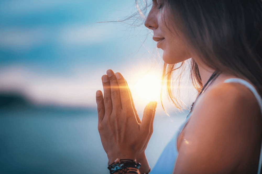 A woman in profile with her eyes closed, holding her hands in a prayer position, with sunlight between her fingers against a background of a blurred ocean and sky.