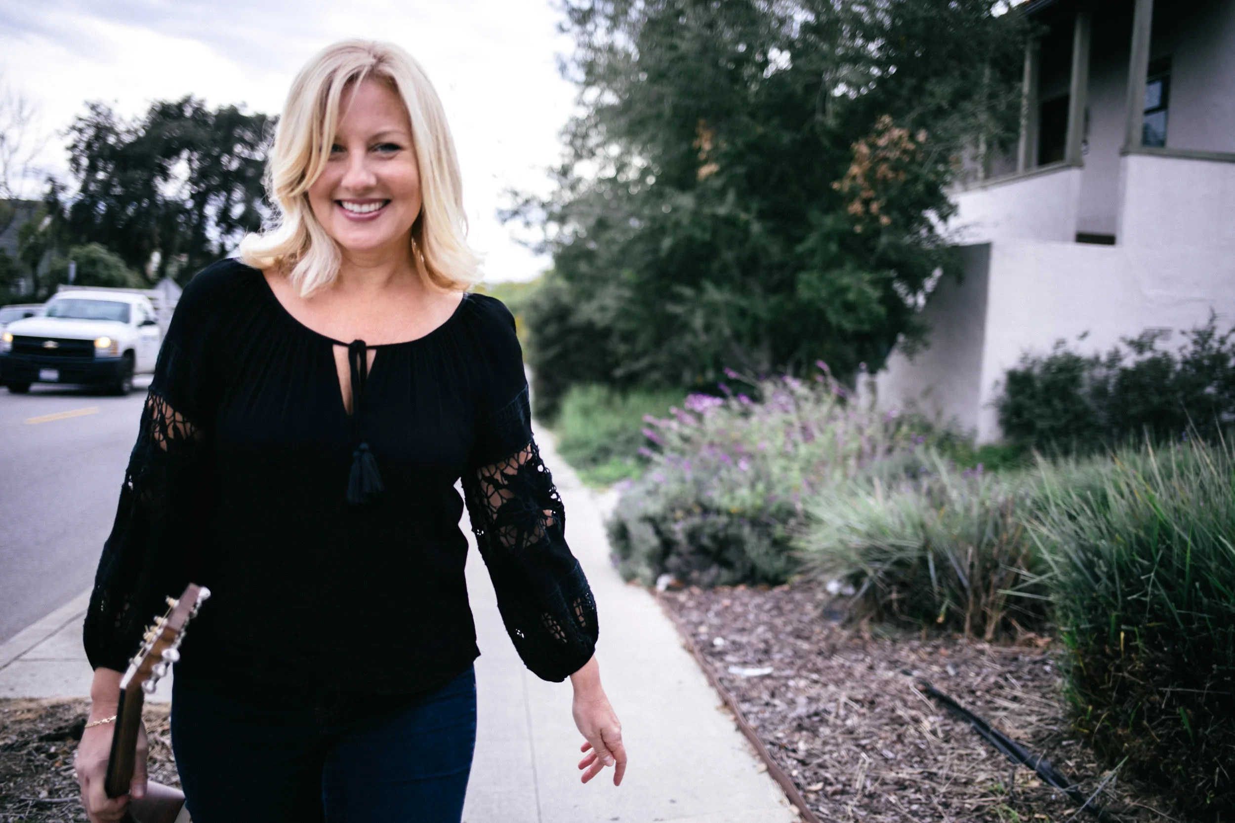 A blonde woman smiling while walking on a sidewalk holding a book and a guitar, with greenery and a white building in the background.