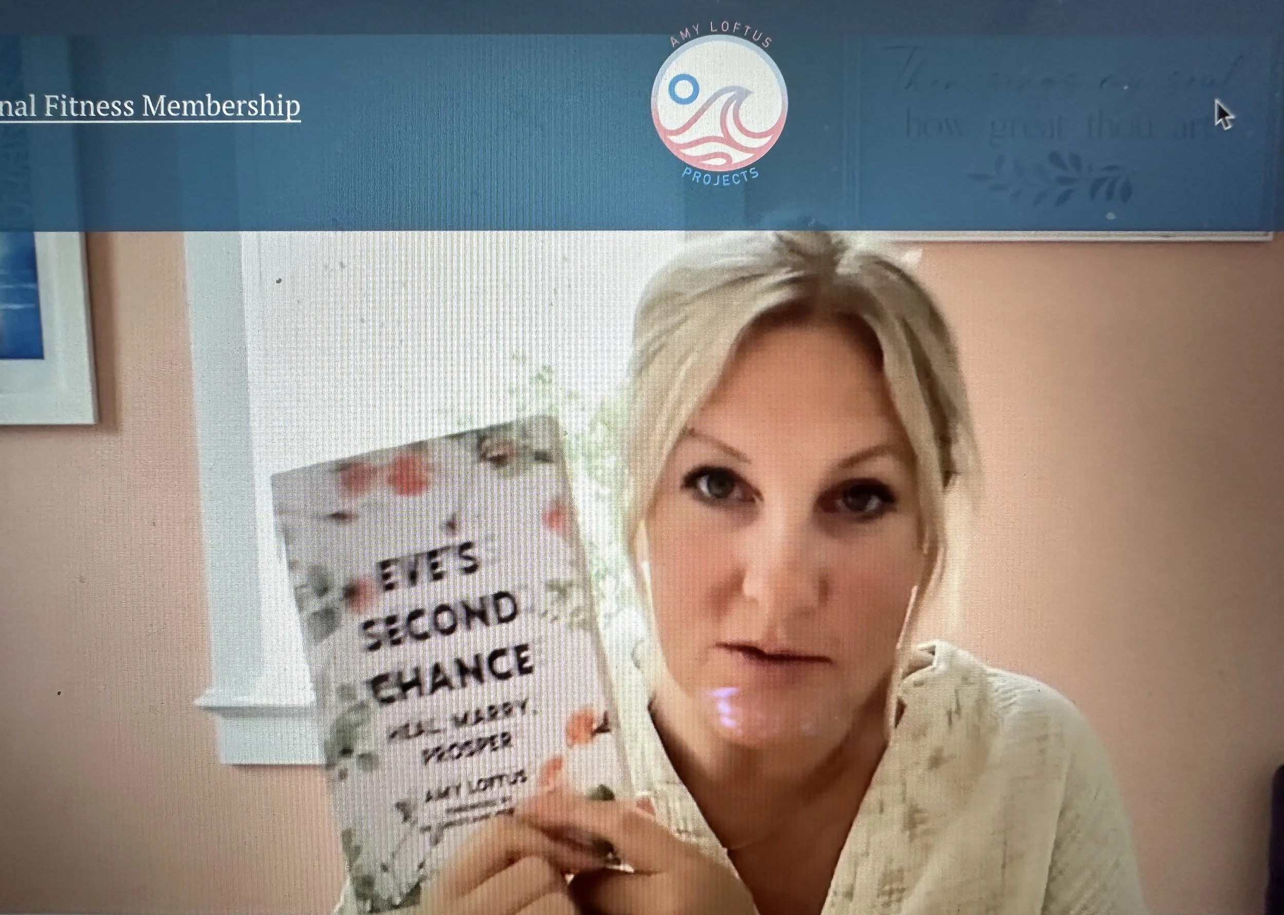 A woman with blonde hair holding a book titled 'It's Second Chance' and looking at the camera. There is a logo at the top with the text 'Amy Loftus Projects' and part of a banner or header visible.