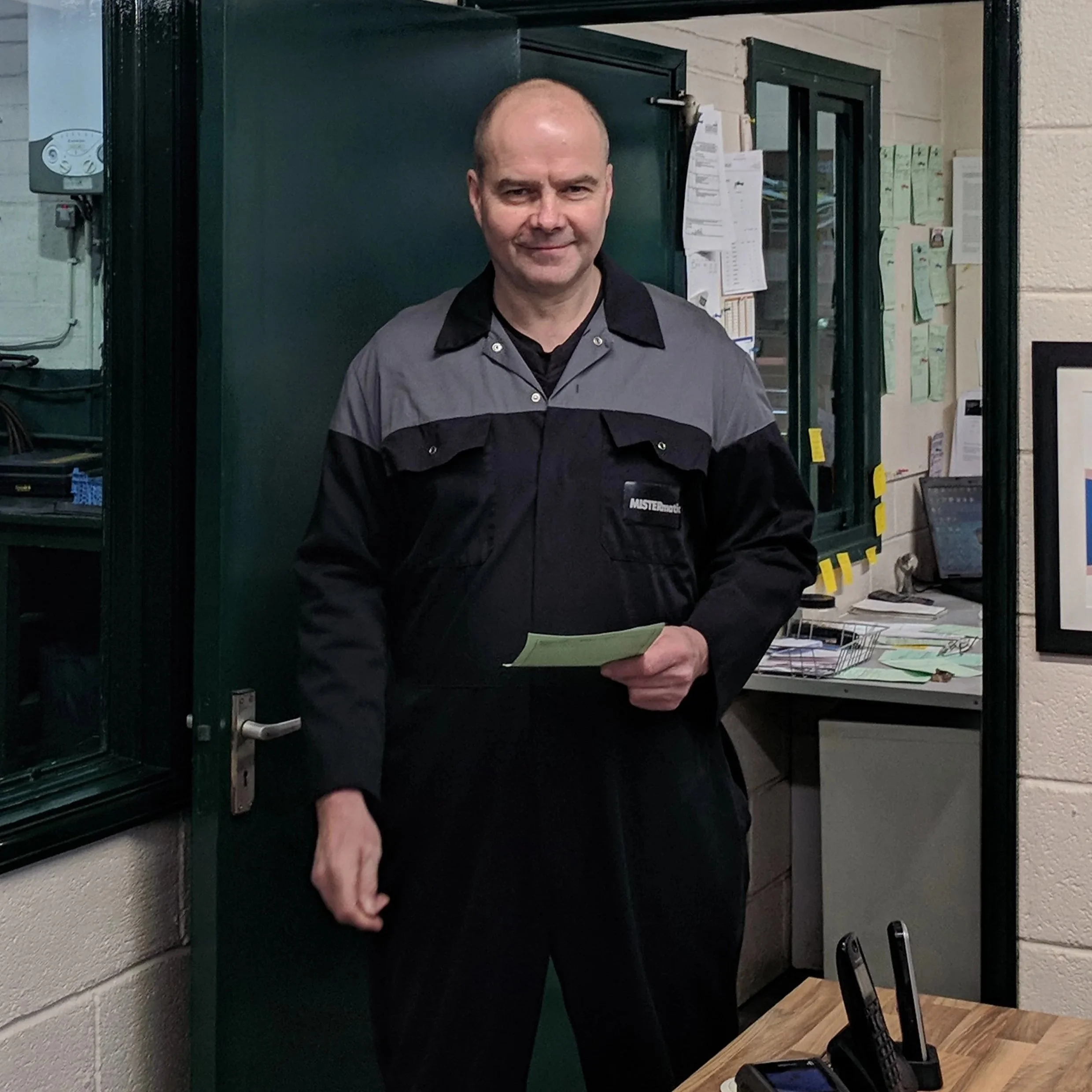 A man in a uniform standing indoors in front of a green door with papers and a bulletin board on the wall behind him.