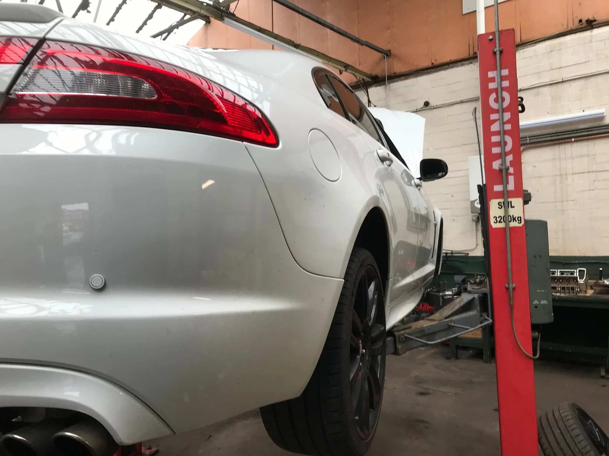 A silver car on a hydraulic lift inside a garage, with a red bar indicating weight capacity of 3200kg, in front of a brick wall with a sign that reads "LIFT UP H".