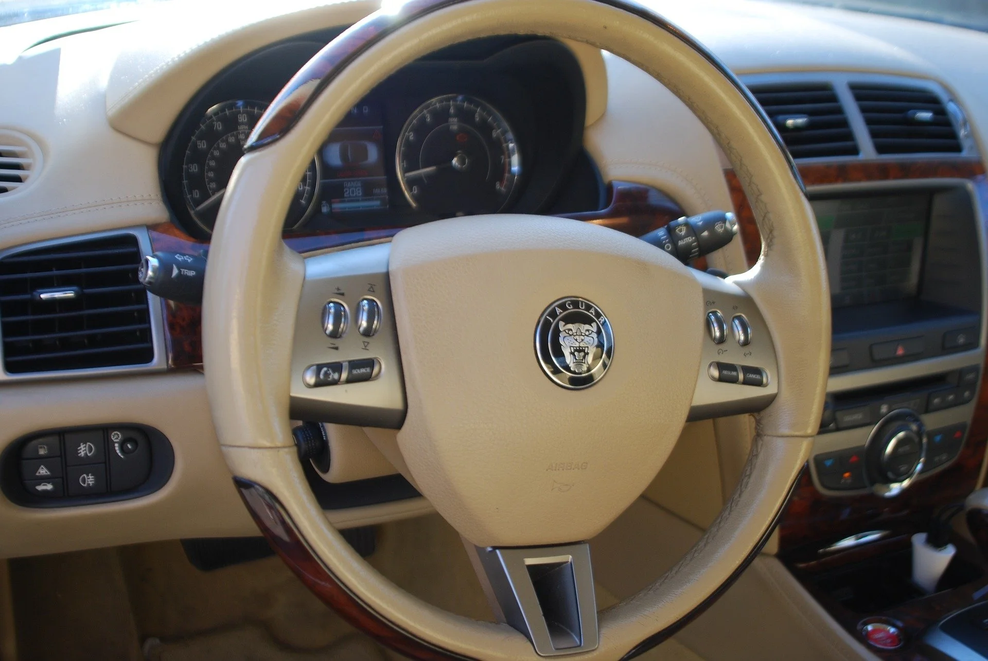 Interior of a luxury car with a beige steering wheel featuring the Jaguar logo, dashboard with speedometer and tachometer, and navigation system.