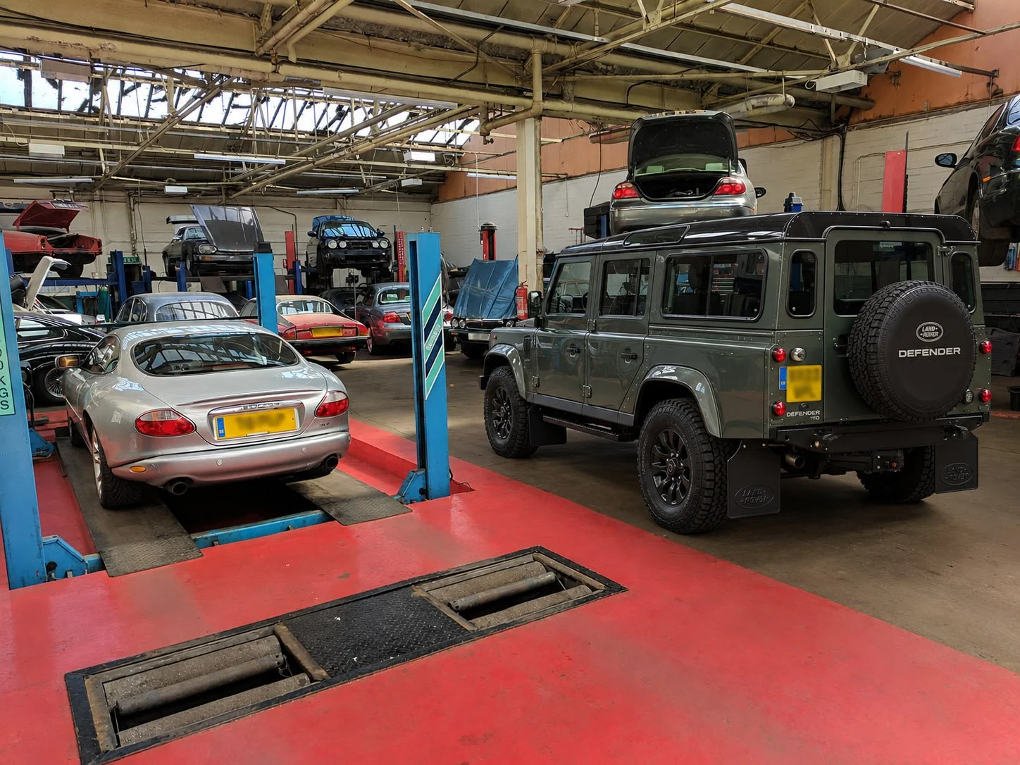 Indoor garage or workshop with multiple cars, including a silver sports car on a hydraulic lift, a green Land Rover Defender, and several other cars on lifts with hoods open.