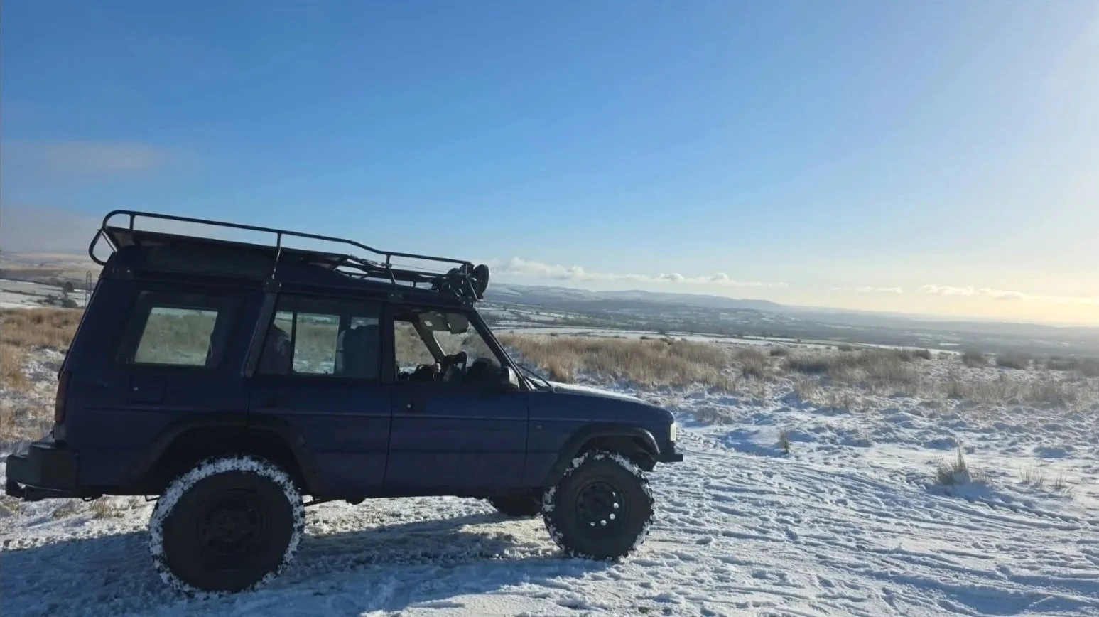 A dark-colored off-road vehicle parked on snow-covered ground in a wide open field under a clear blue sky, with distant hills and clouds on the horizon.
