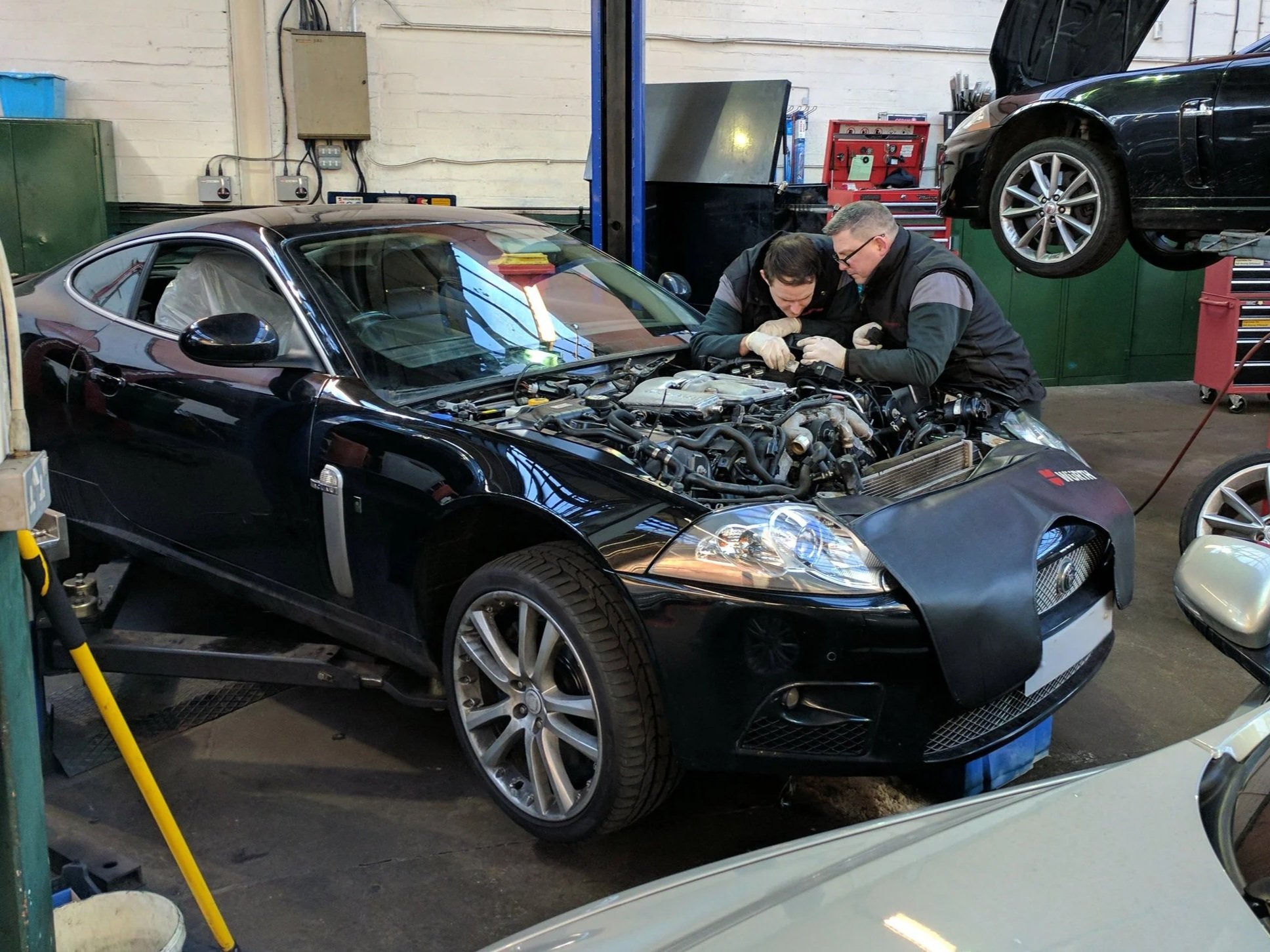 Two mechanics working on a black sports car with its hood open in an auto repair shop, with another black car elevated on a lift in the background.