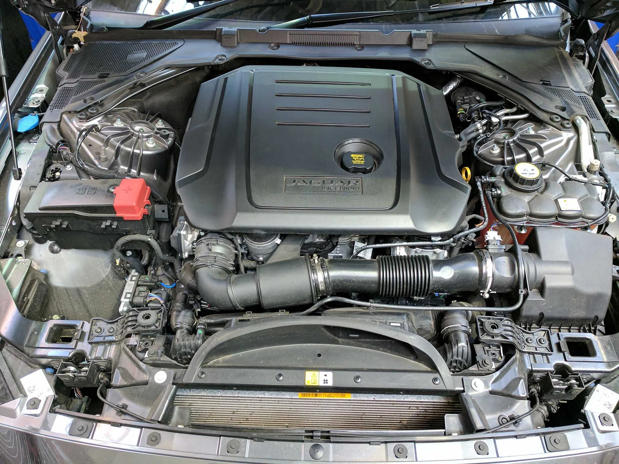Under the hood of a modern Jaguar vehicle, showing the engine with a large black cover labeled 'Jaguar Ingenium' surrounded by various tubes, wires, and mechanical components.