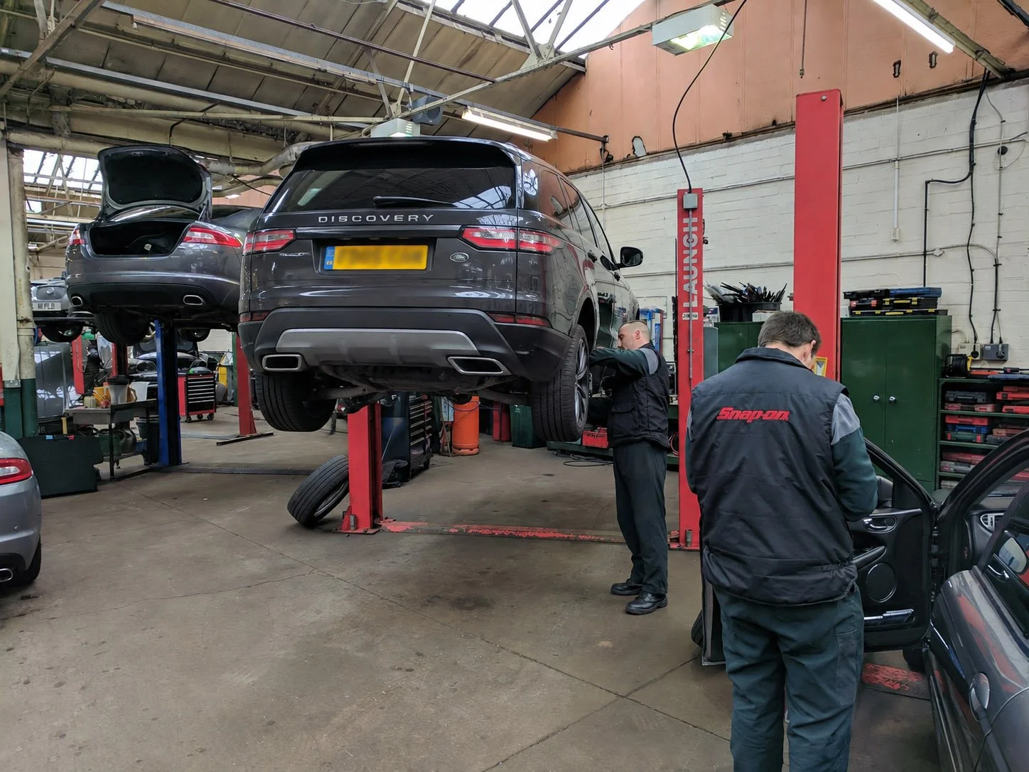 Auto repair shop with a gray Land Rover Discovery elevated on a hydraulic lift. Two mechanics are working on the vehicle; one is inspecting the right rear wheel, and the other is handling tools. The shop has various equipment, tools, and other cars in the background.