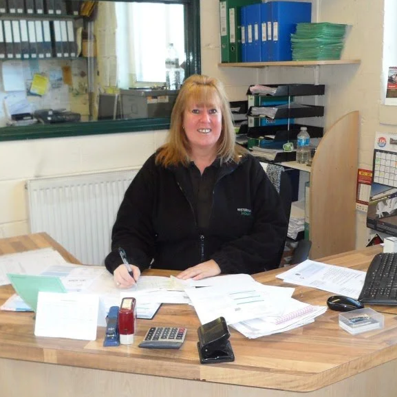 A woman sitting at a cluttered desk in an office, smiling, surrounded by papers, office supplies, and a computer.
