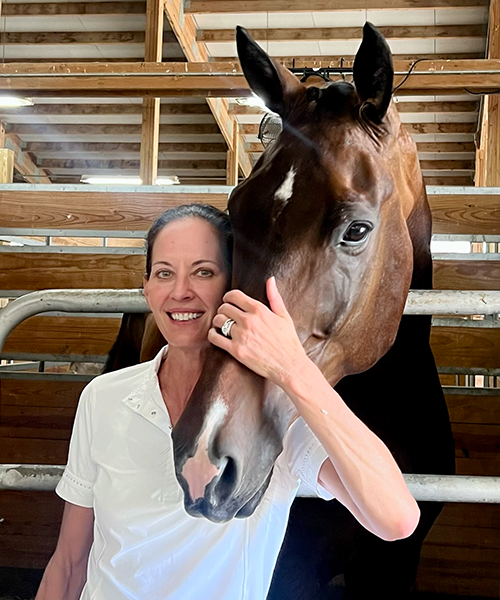 A woman standing next to a brown horse inside a wooden stable, with the woman smiling and the horse's head close to her face.
