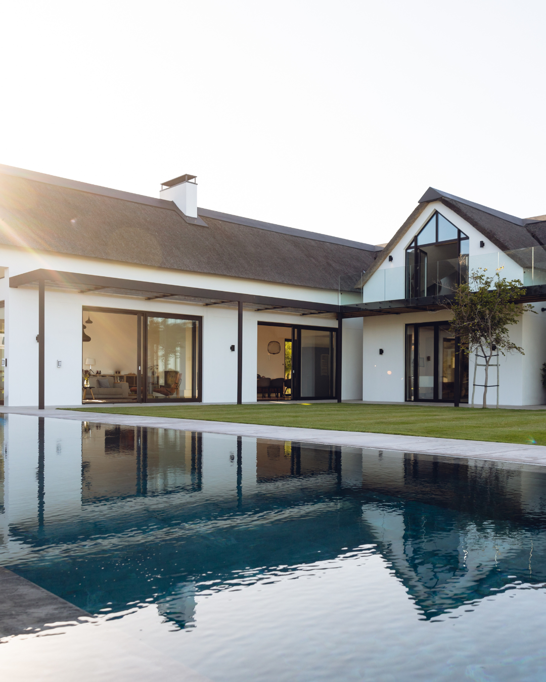 Modern house with white exterior, large glass windows, a swimming pool in the foreground, and a well-maintained lawn, during daytime.