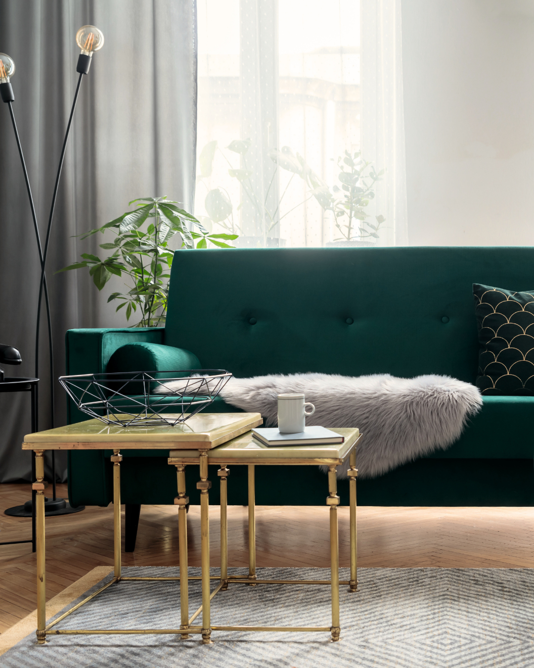Living room with green sofa, gold side tables, a white rug, a black throw pillow, a mug, books, a faux fur blanket, potted plants, a window with sheer curtains, and a tall black floor lamp.