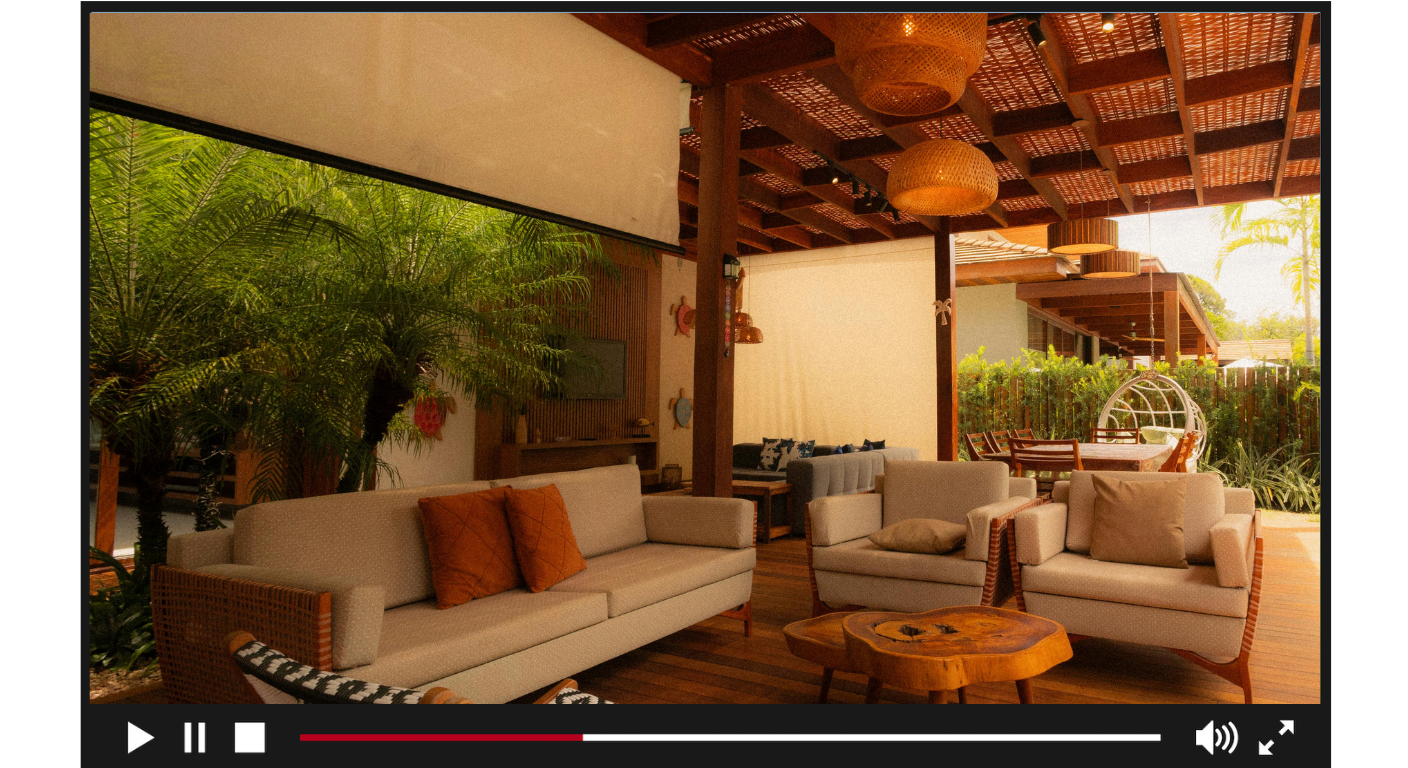 A cozy outdoor patio with wooden furniture and white sofas surrounded by lush green plants and trees, featuring a wooden ceiling with hanging wicker pendant lights.