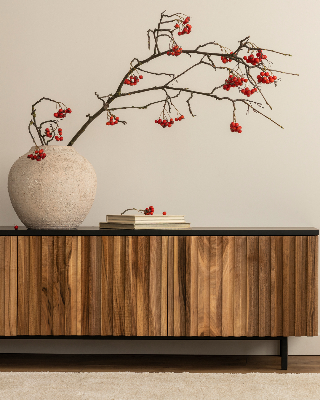 A large off-white ceramic vase with a textured surface holding a bare branch with red berries. The branch extends upward and outward, with some berries resting on a closed book or magazine on a black-topped wooden sideboard against a plain wall.