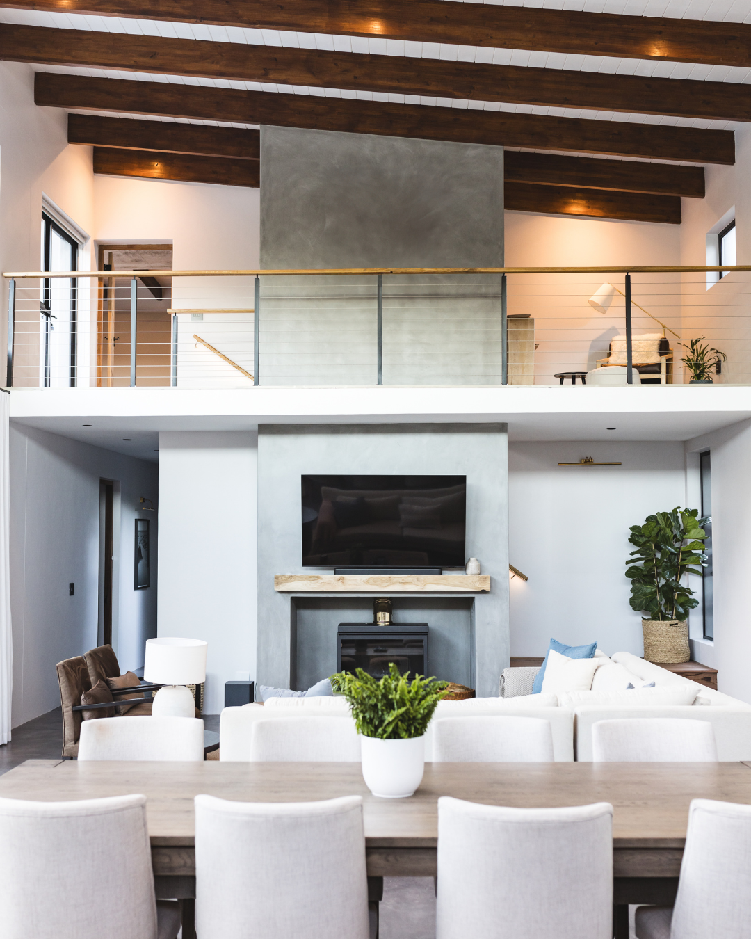 Modern living room with a dining table in the foreground, a fireplace with a mounted TV above it, a white sofa, and an upstairs loft with seating and plants, featuring wood-beamed ceilings.