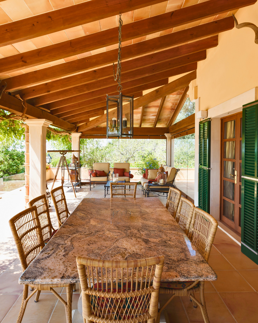 Covered outdoor patio with a large marble dining table, wicker chairs, and a seating area with cushioned chairs and a small table. The ceiling has exposed wooden beams and a lantern-style chandelier. There are green shutters on the wall and lush greenery visible in the background.