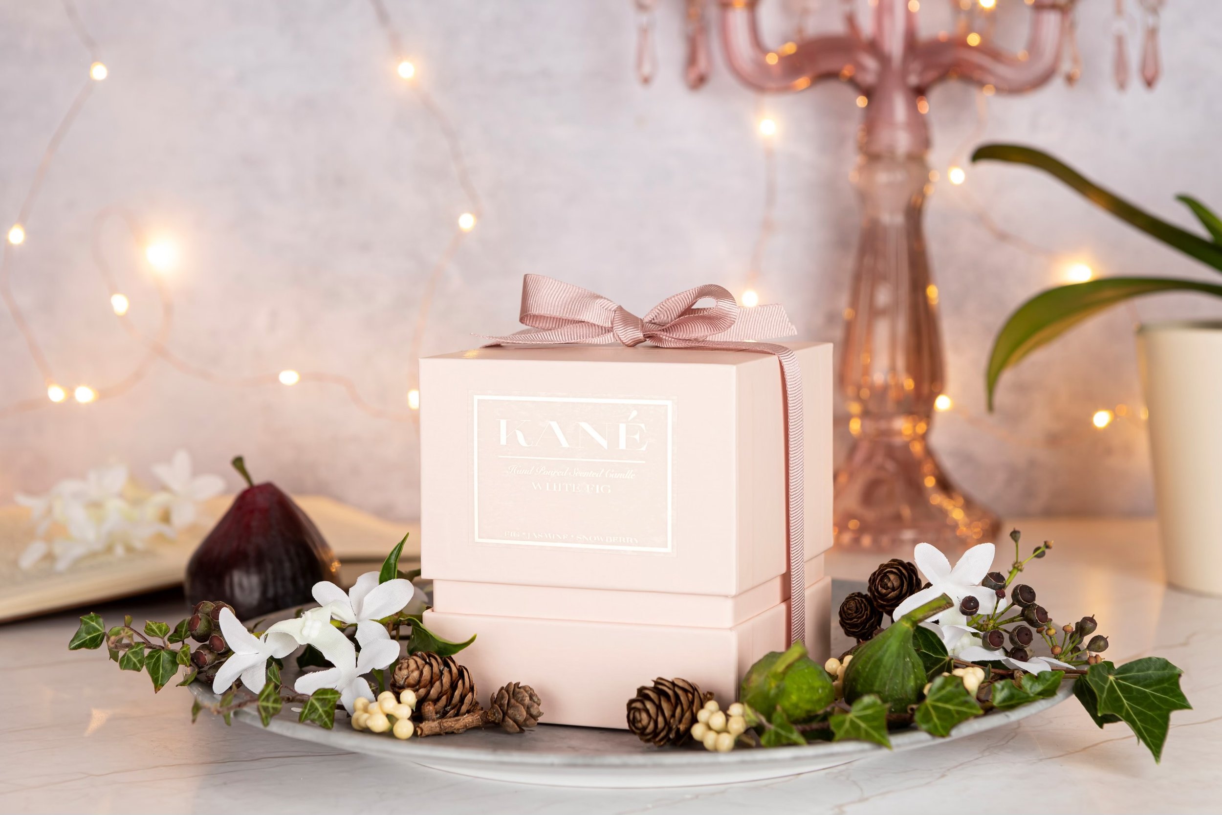 Decorative table setting with a pink gift box, surrounded by white flowers, greenery, pinecones, and berries, with a rose-colored chandelier and fairy lights in the background.