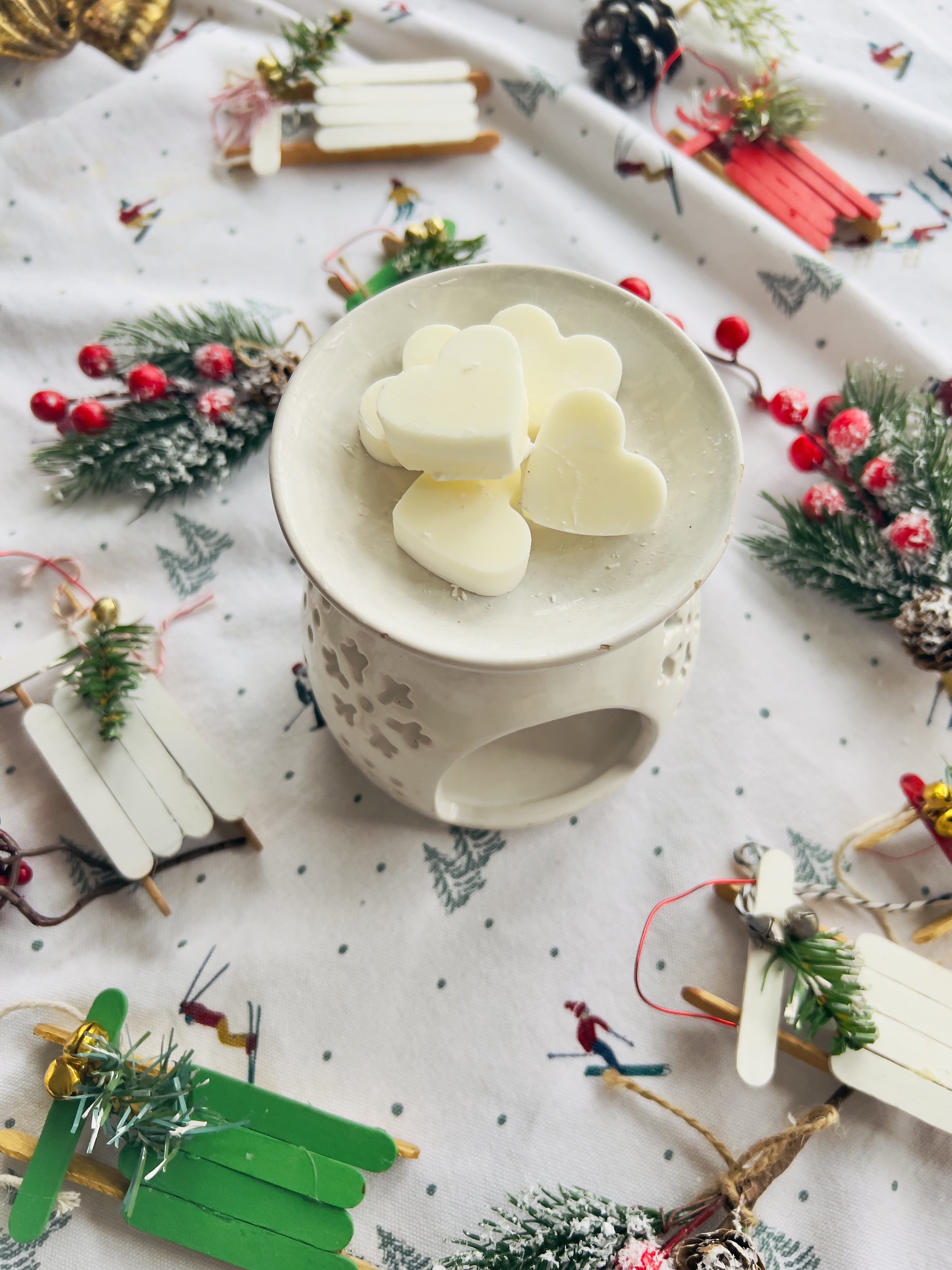 White candle holder with heart-shaped white candles on top, decorated with small Christmas-themed greenery, on a holiday tablecloth with Christmas imagery and tiny decorated Christmas trees.