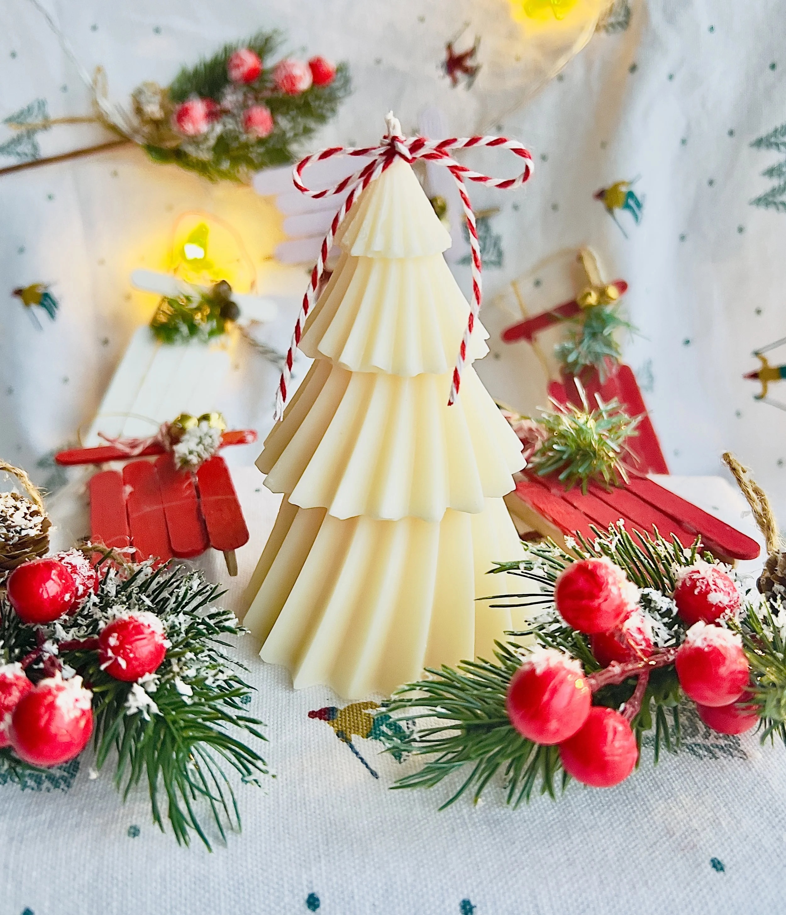 Decorative Christmas scene with a white chocolate Christmas tree tied with red and white string, surrounded by holly with red berries, red miniature sled, and festive background.