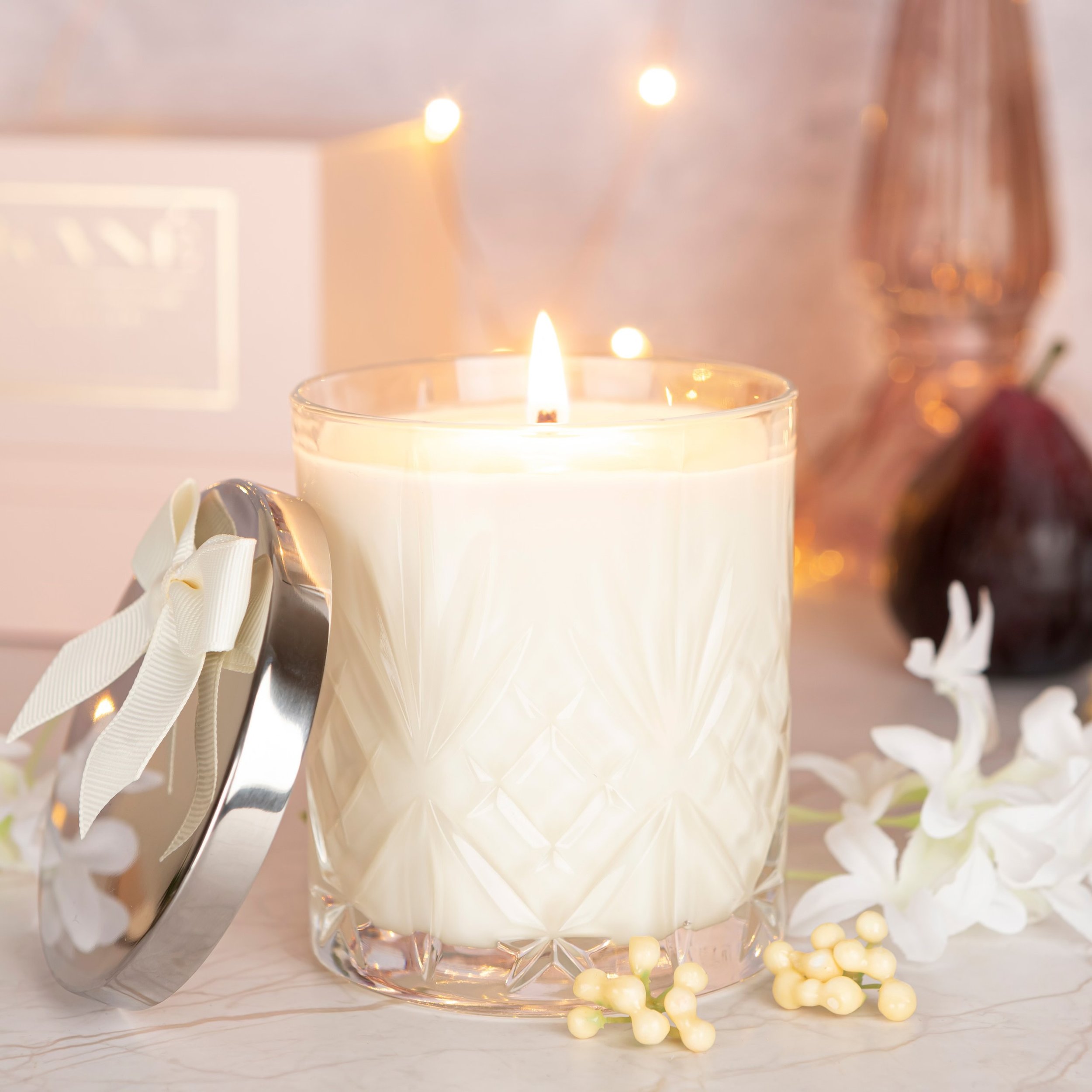 A lit candle in a decorative crystal glass holder with a metal lid decorated with a white ribbon bow. White flowers and yellow berries are placed around it.
