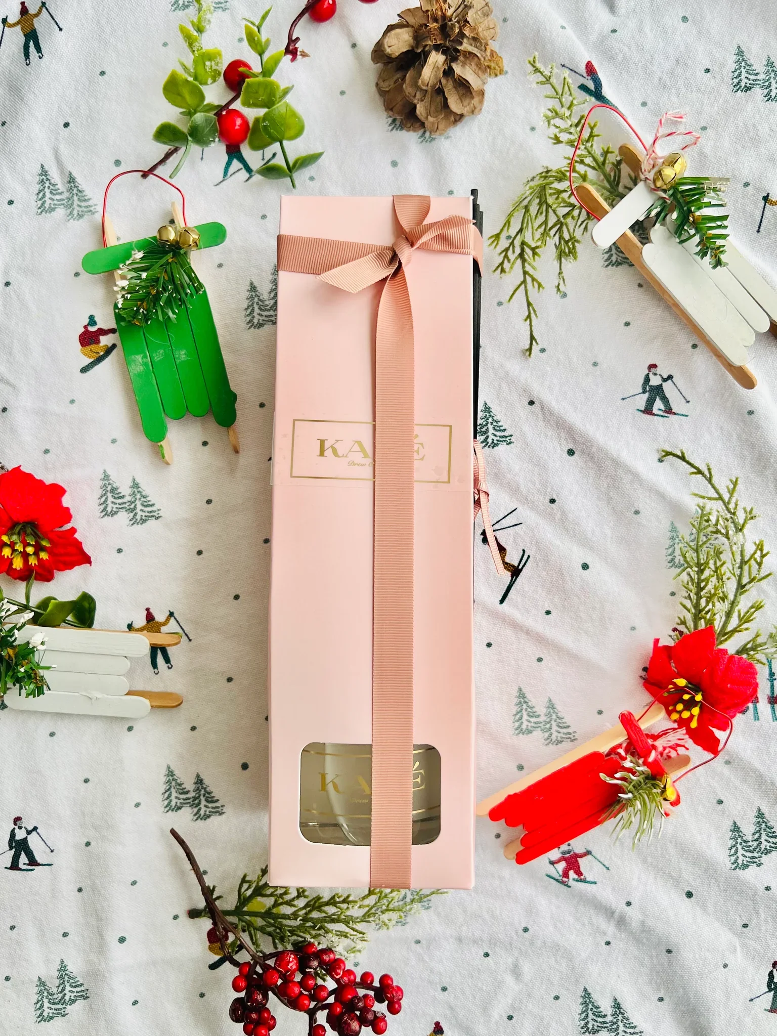Pink gift box with a ribbon bow on a Christmas-themed tablecloth surrounded by small holiday crafts and decorations.