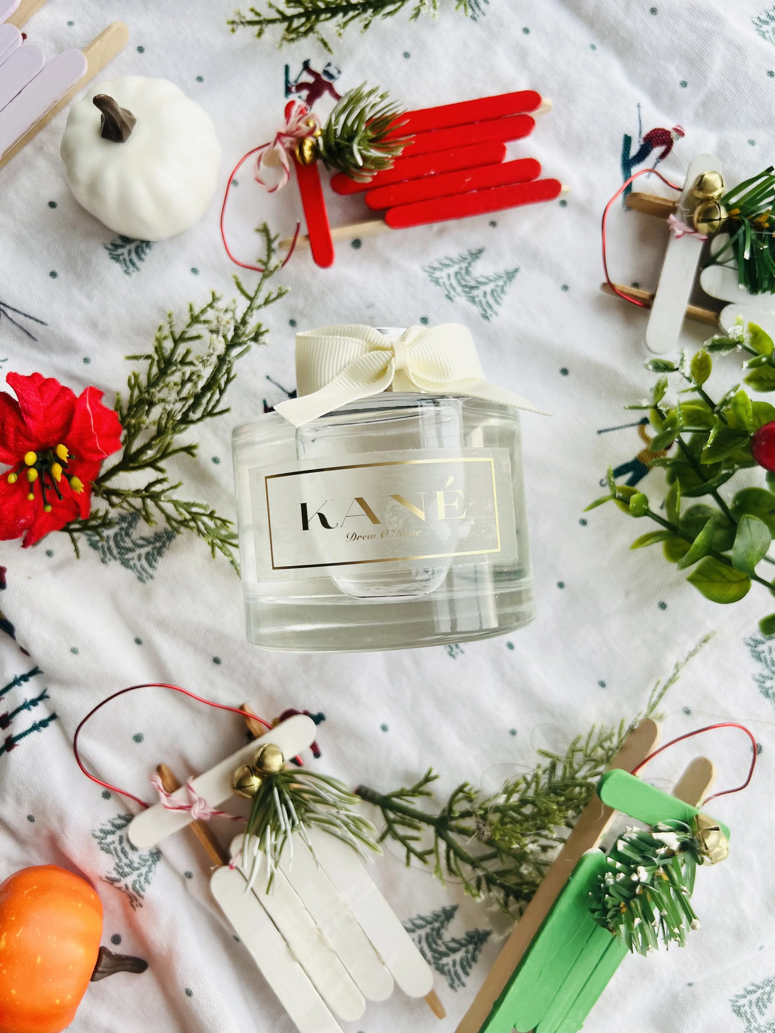 Holiday-themed arrangement with a glass perfume bottle labeled 'KANE' with a white ribbon bow, surrounded by miniature decorative pumpkins, small flowers, greenery, and colorful popsicle stick sleighs on festive tablecloth.