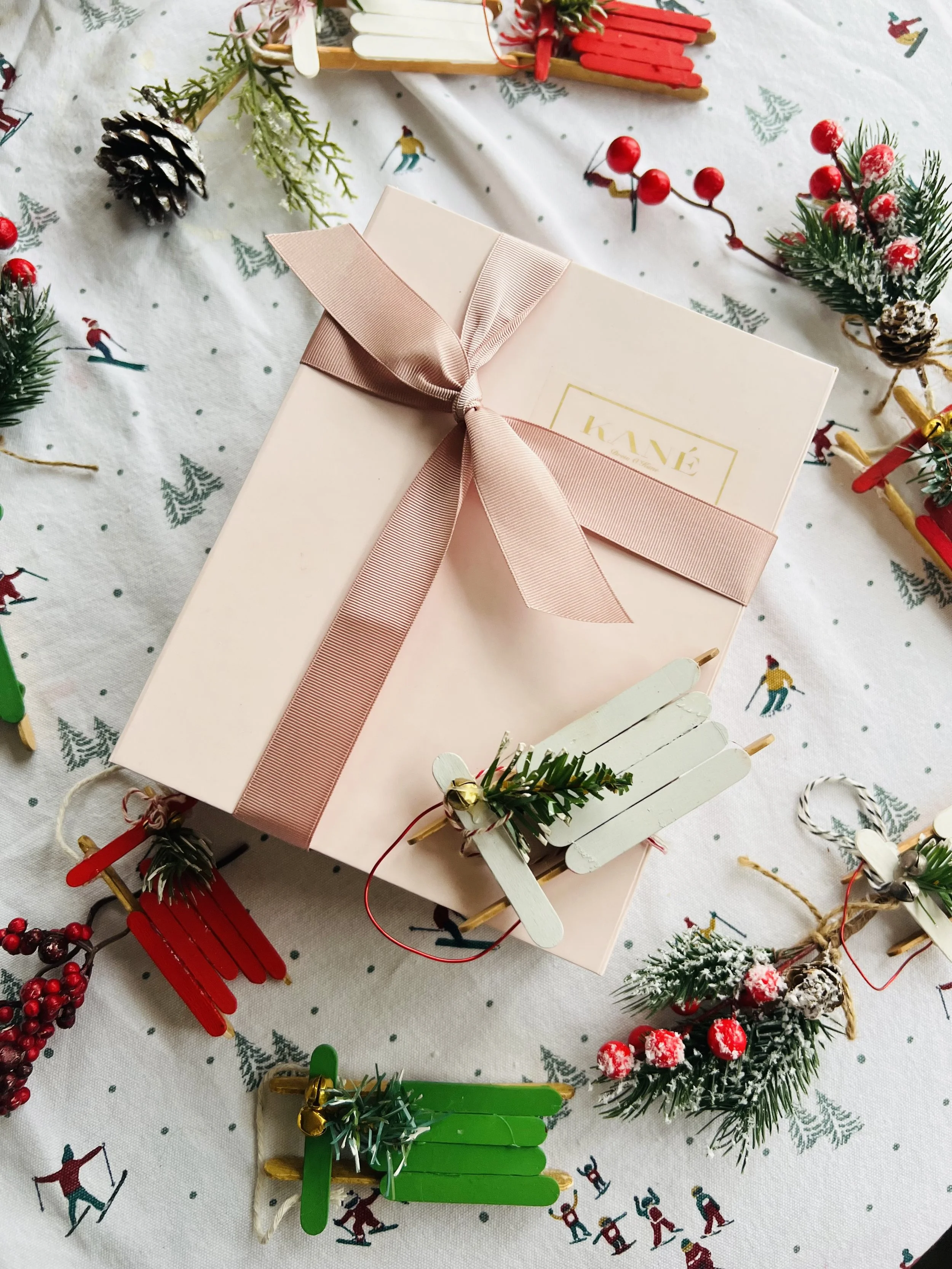 Christmas gift box wrapped with pink paper and a pink ribbon bow, surrounded by miniature sleds, pinecones, and festive decorations on a tablecloth with a winter theme.