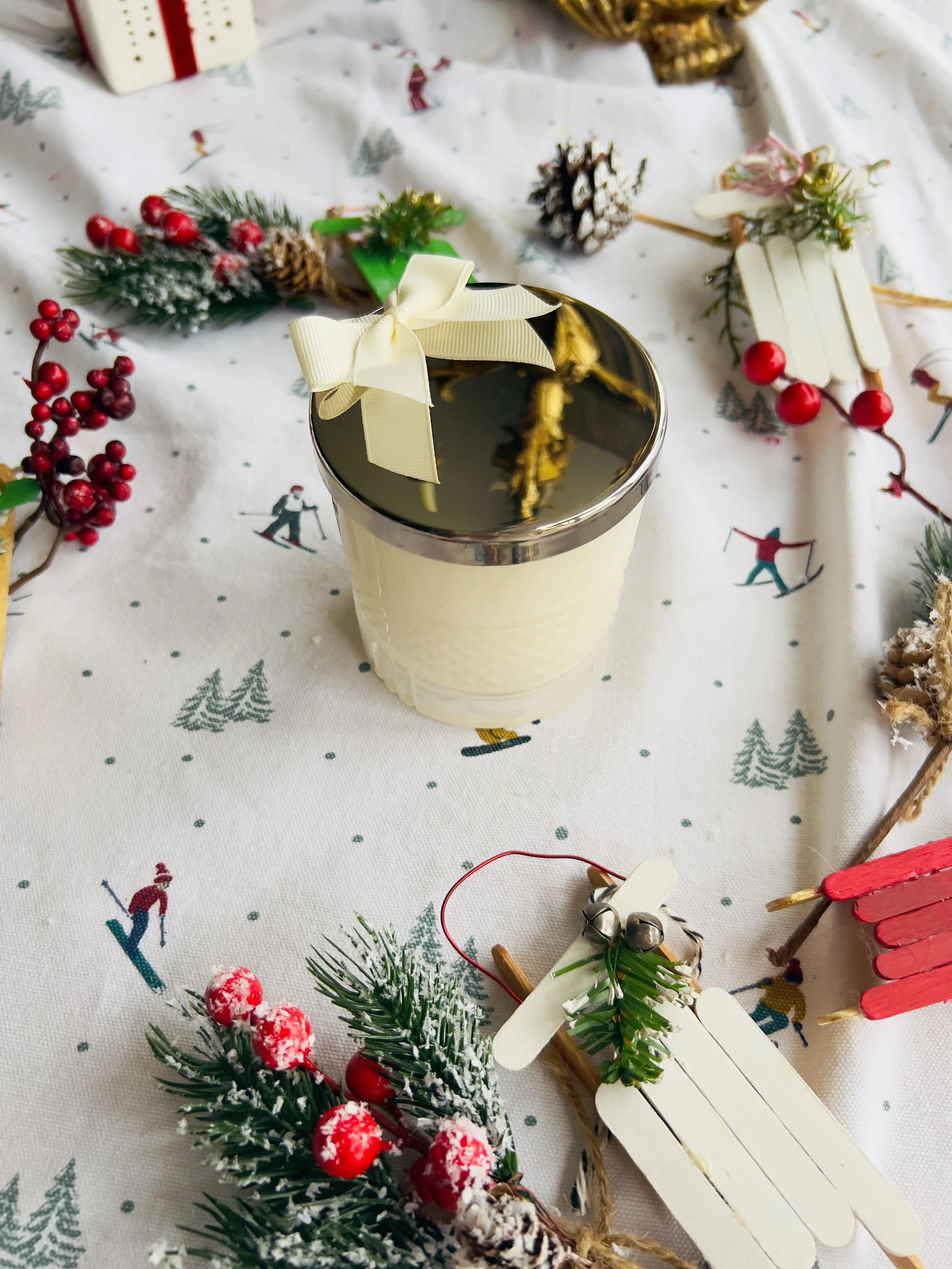 A glass of dark beverage with a cream-colored bow on top, surrounded by Christmas decorations, pinecones, faux snow, and festive figurines on a holiday-themed tablecloth featuring skiers and pine trees.