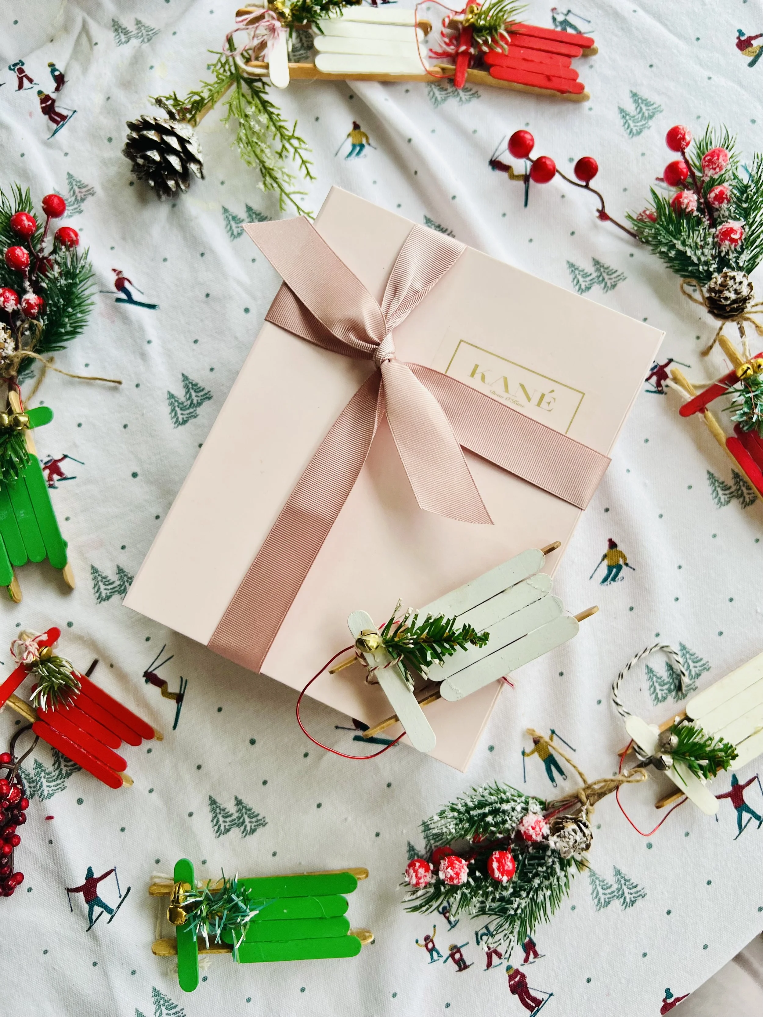A pink gift box wrapped with a pink ribbon, placed on a festive tablecloth decorated with holiday-themed drawings and surrounded by Christmas ornaments and decorations, including miniature sleds, pinecones, berries, and greenery.
