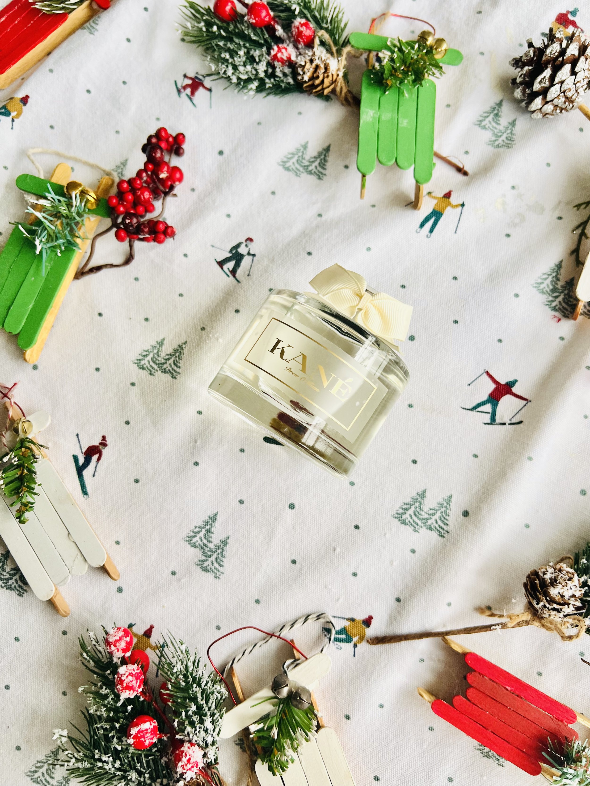 A set of holiday decorations on a white Christmas-themed tablecloth with small trees and skiers. Centered is a clear perfume bottle labeled 'KANE' with a cream-colored ribbon bow on top.