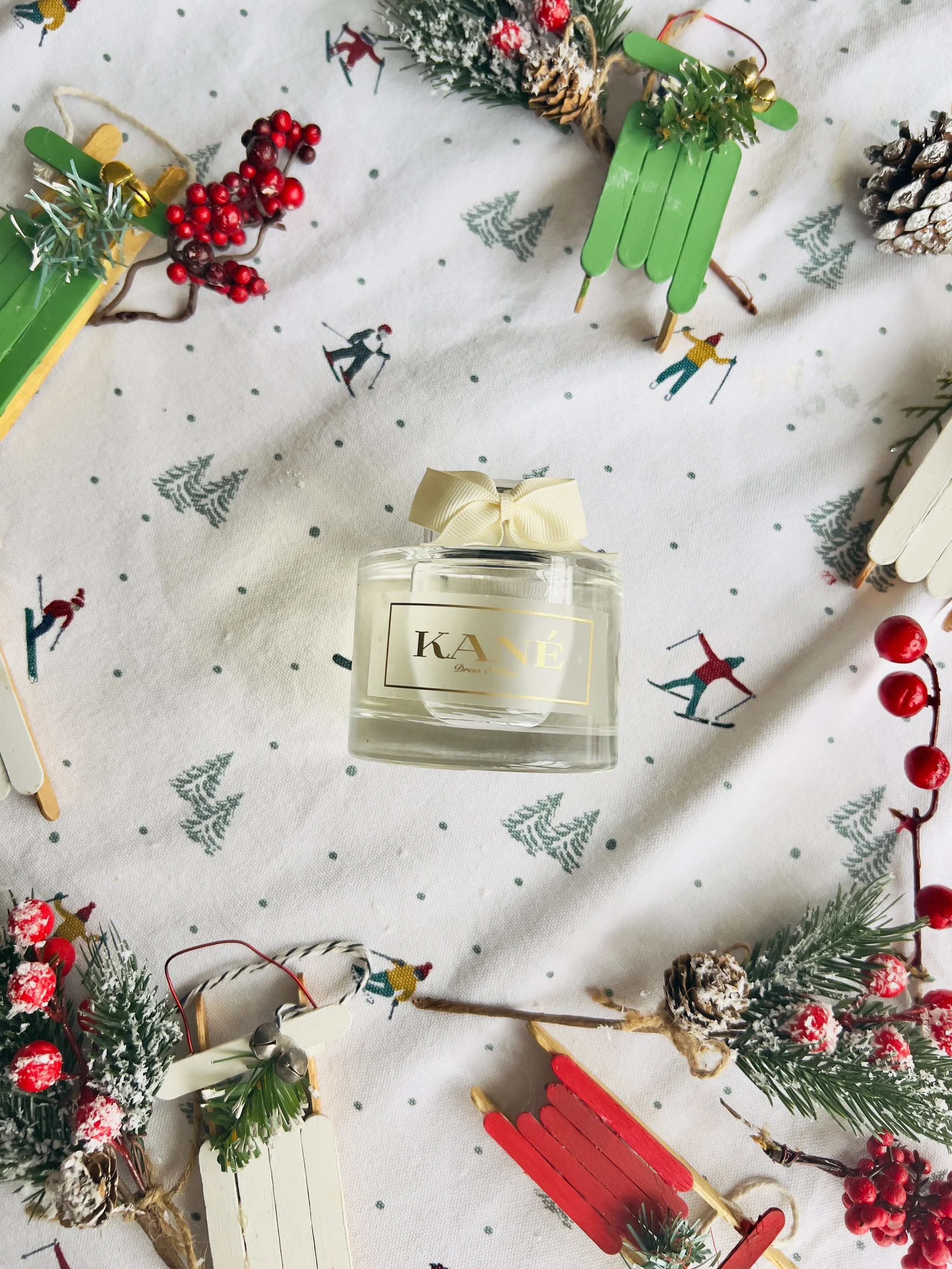 A glass jar of KANE perfume with a cream-colored bow on top, placed on a white cloth decorated with small trees and skiers, surrounded by holiday decorations including miniature sleds, pinecones, red berries, and sprigs of greenery.