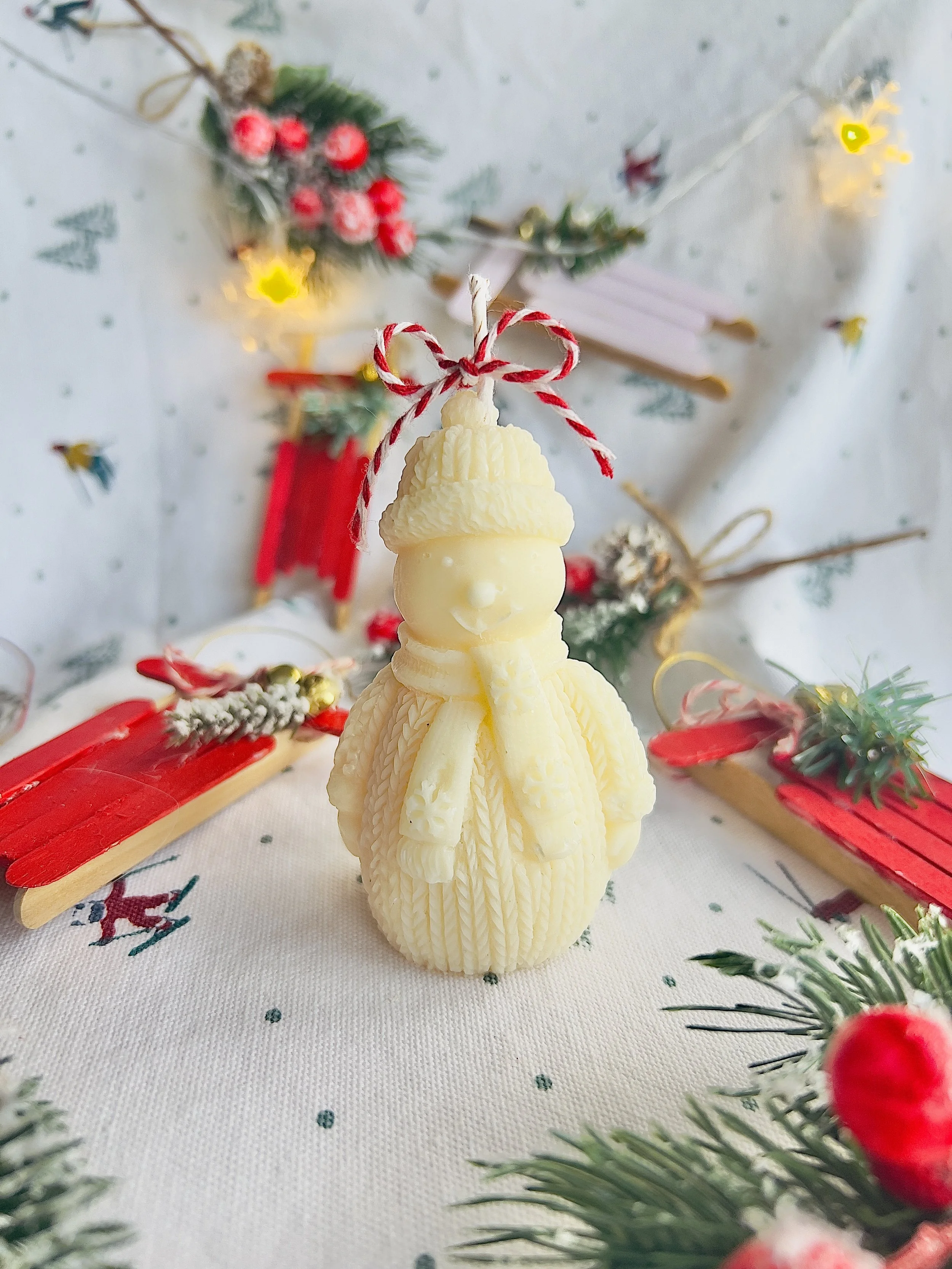 A white snowman-shaped candle with a textured hat and scarf, decorated with a red and white striped bow on top, surrounded by Christmas decorations including red and white sleds, greenery, berries, and fairy lights, on a festive tablecloth.