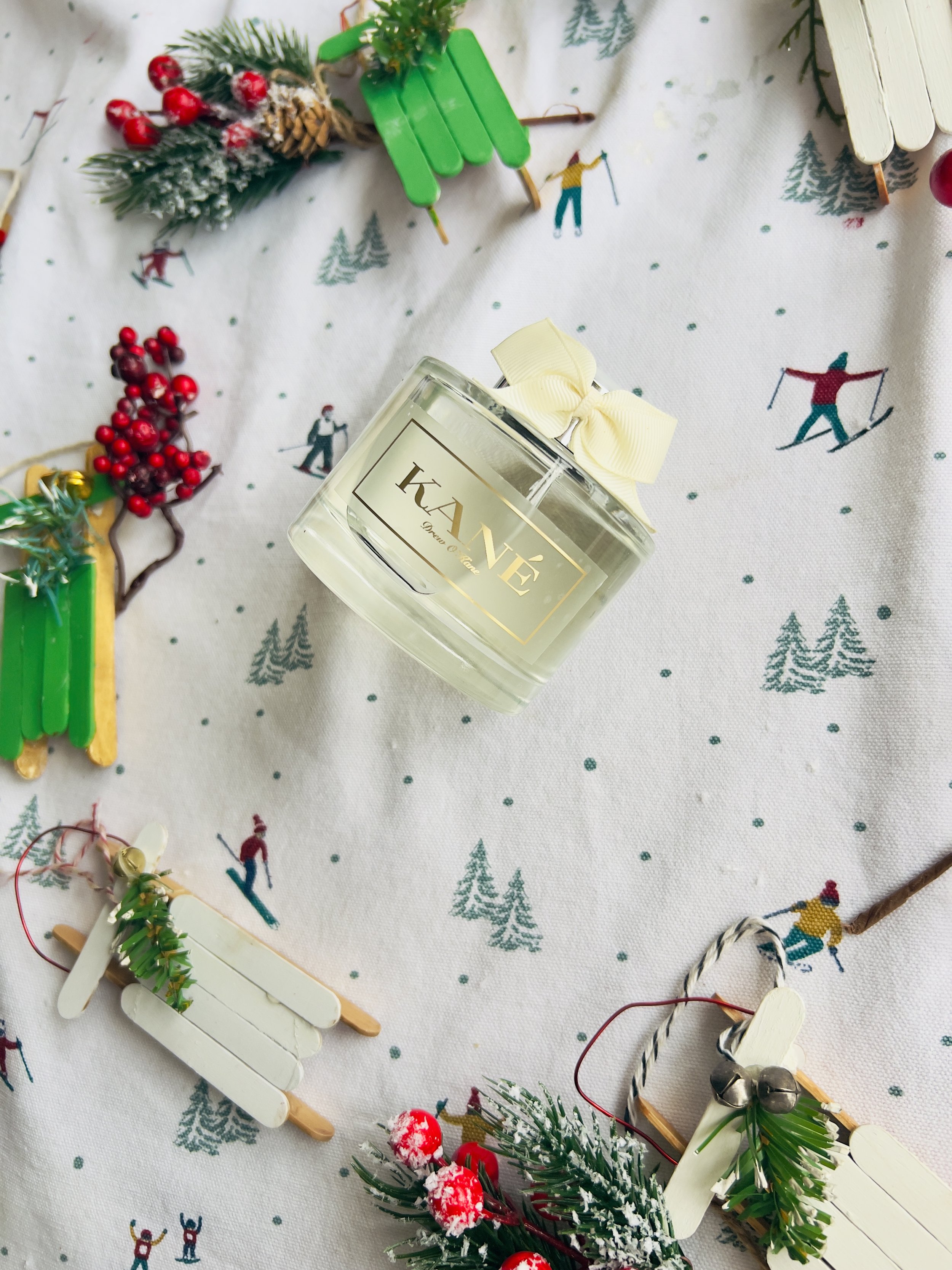 A glass candle jar with a yellow bow sits on a festive Christmas-themed tablecloth, surrounded by miniature sled decorations adorned with greenery and red berries.