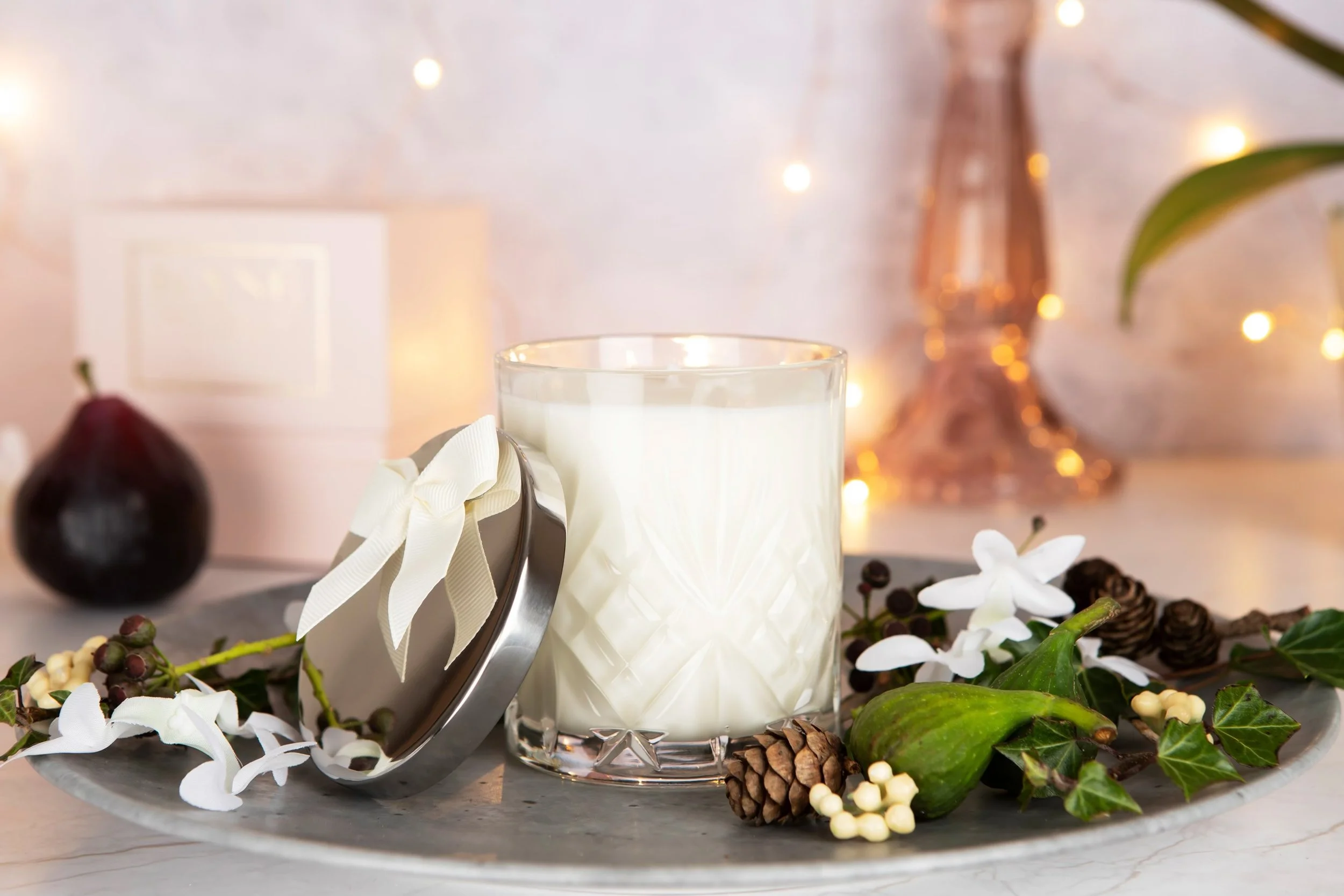 A glass of white candle decorated with Christmas greenery, pinecones, and flowers on a silver tray, with a blurred background of fairy lights and a fig in the background.