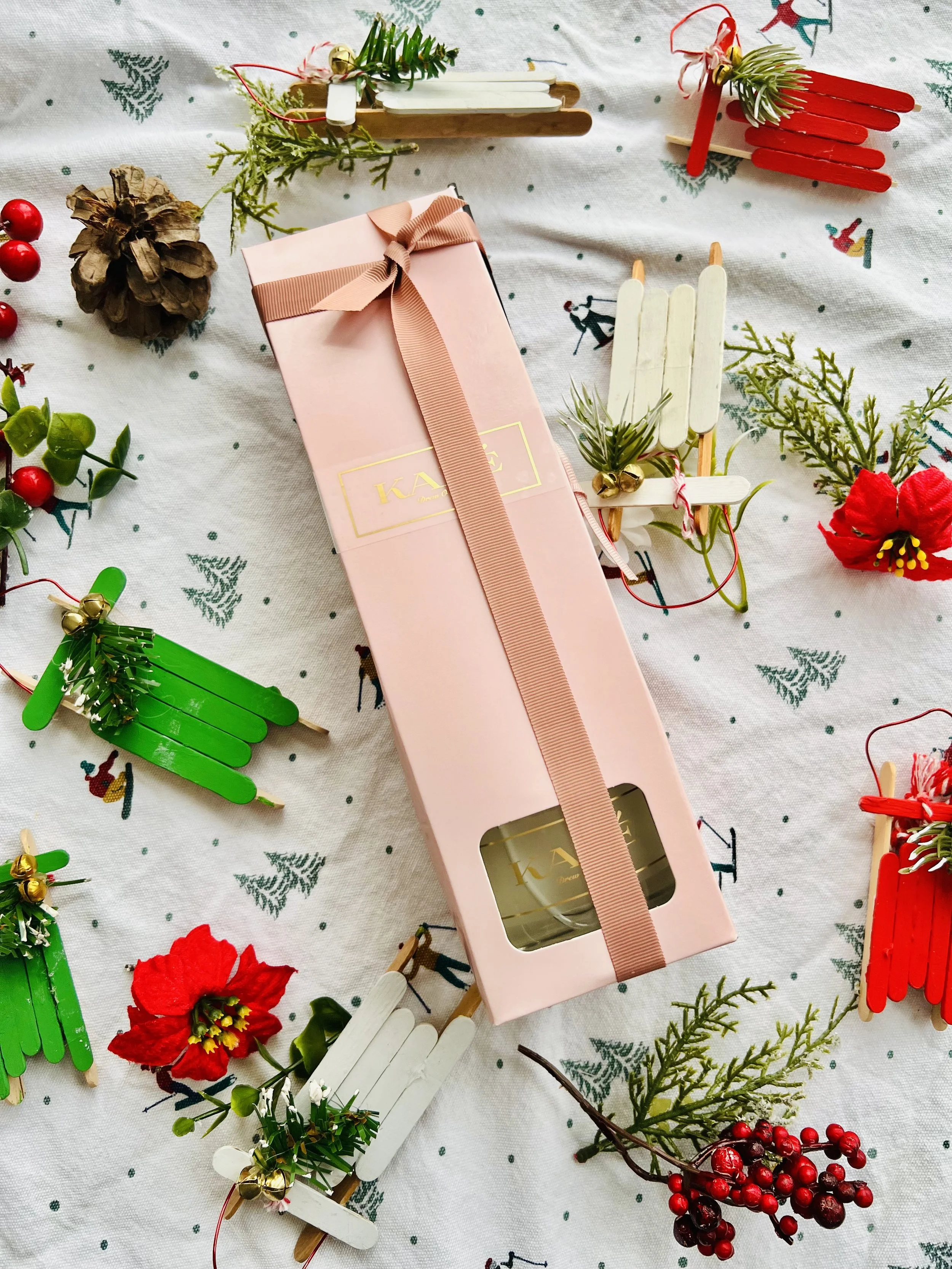 Pink gift box with a pink ribbon, surrounded by Christmas-themed decorations including red and green ornaments, artificial flowers, small decorative sleds, sprigs of greenery, and berries on a festive cloth.