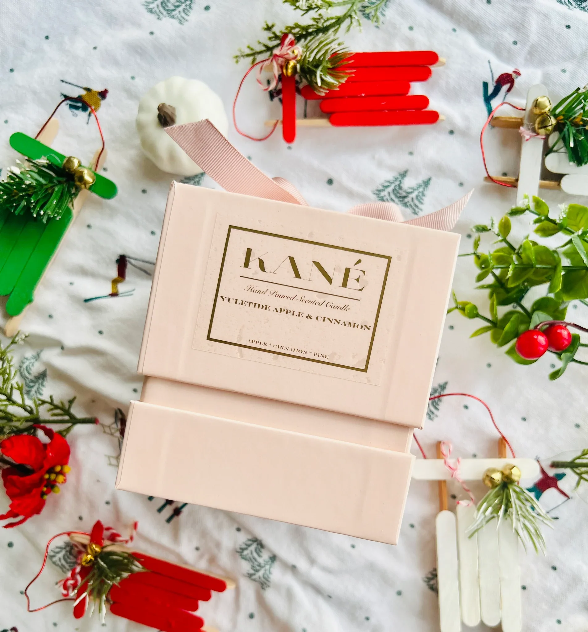 A beautiful pink box wrapped in pink ribbon with a gold label named KANÉ scented candle with apple and cinnamon scent, surrounded by holiday decorations including miniature sleds, sprigs of greenery, and red berries on a white festive tablecloth.