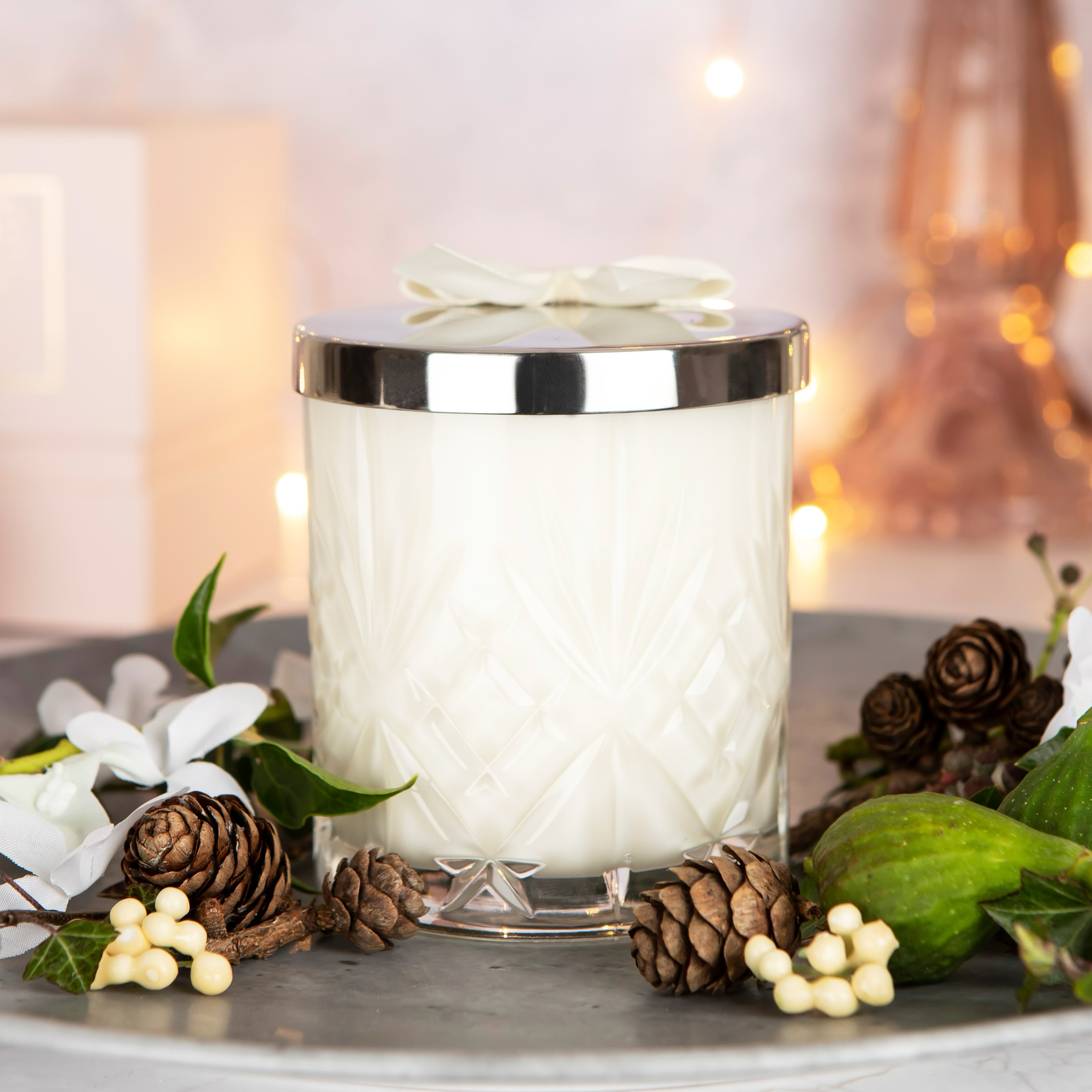A decorative white candle in a textured glass holder with a silver lid and bow on top, surrounded by pinecones, green leaves, white berries, and a green fruit; blurred warm background with lights.