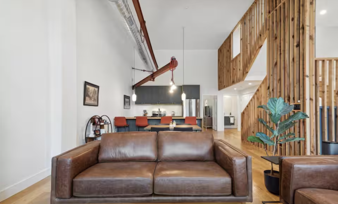 Interior of a modern living space featuring a leather sofa, a wooden staircase with a railing, a plant in a pot, a dining area with orange chairs, and a kitchen with a black backsplash and stainless steel appliances.