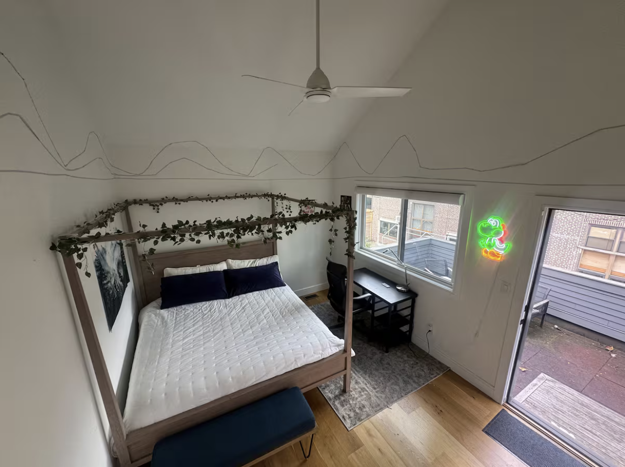 A bedroom with a wooden bed frame decorated with fake vines, a white quilt, and dark pillows. There is a black desk with a chair under a window, a neon sign of a green cartoon frog on the wall, and a ceiling fan. The room has hardwood floors and an open door leading to a small outdoor patio.