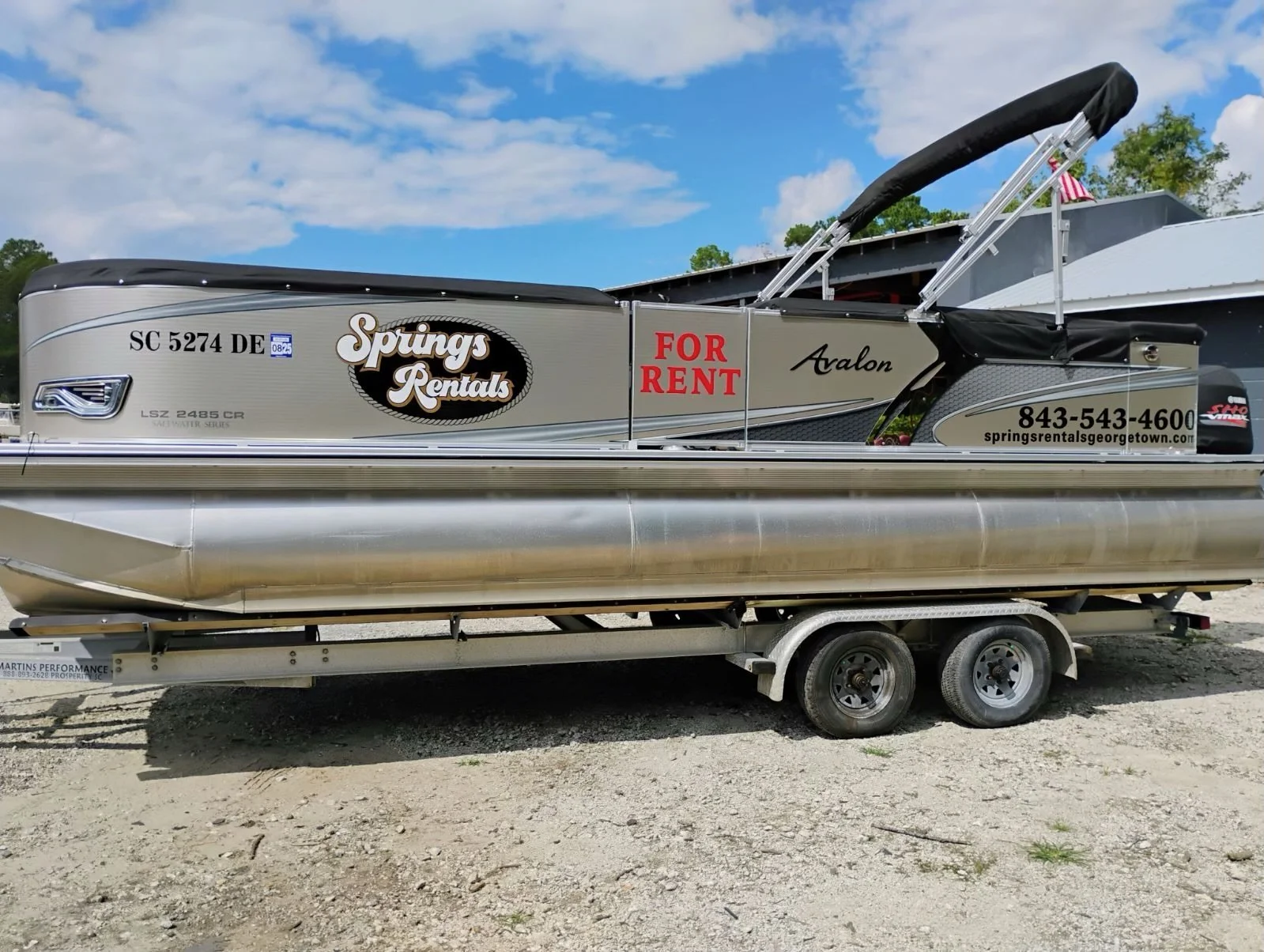 A pontoon boat on a trailer with advertising for Spring's Rentals, parked on gravel with blue sky and clouds in the background.