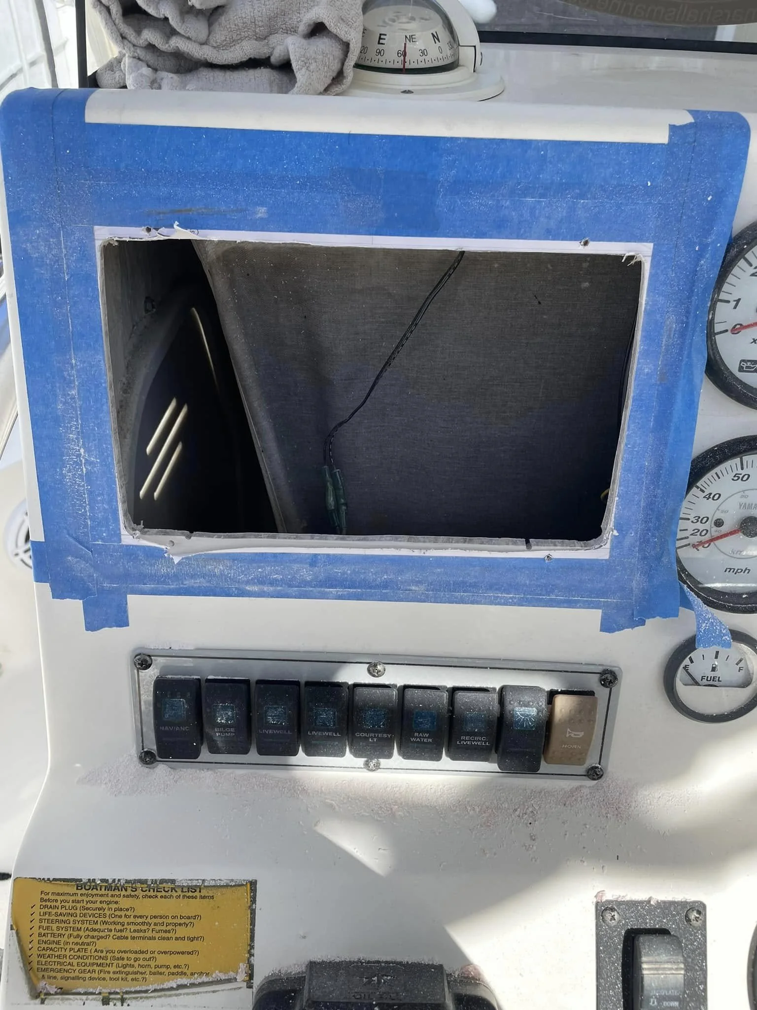 The control panel of a boat's dashboard shows a rectangular opening with no cover, a row of switches labeled for various boat functions, a warning label, and some gauges and a gray cloth on top.