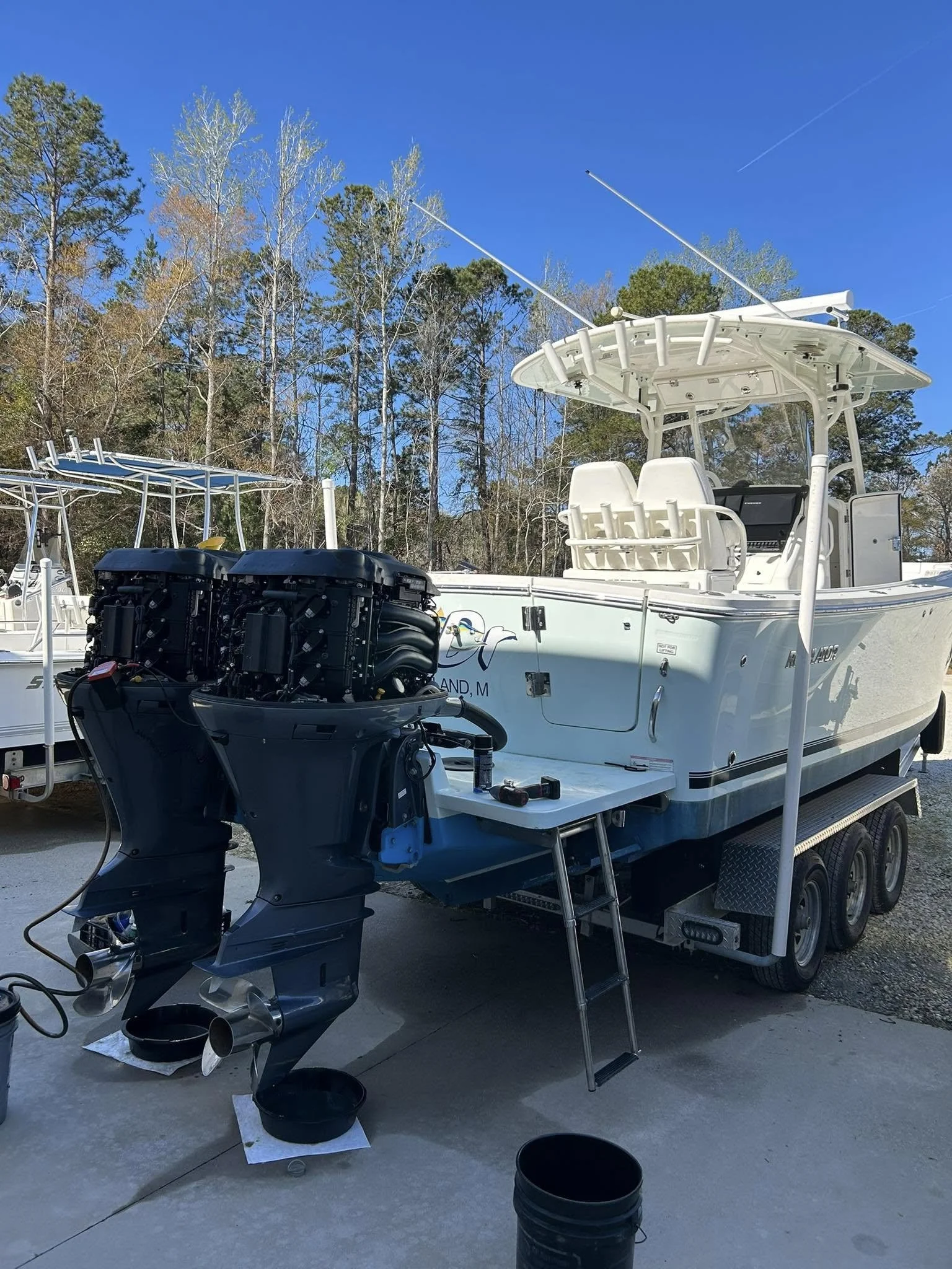A white fishing boat with twin engines on a trailer, parked outdoors on a concrete surface, surrounded by trees against a bright blue sky.