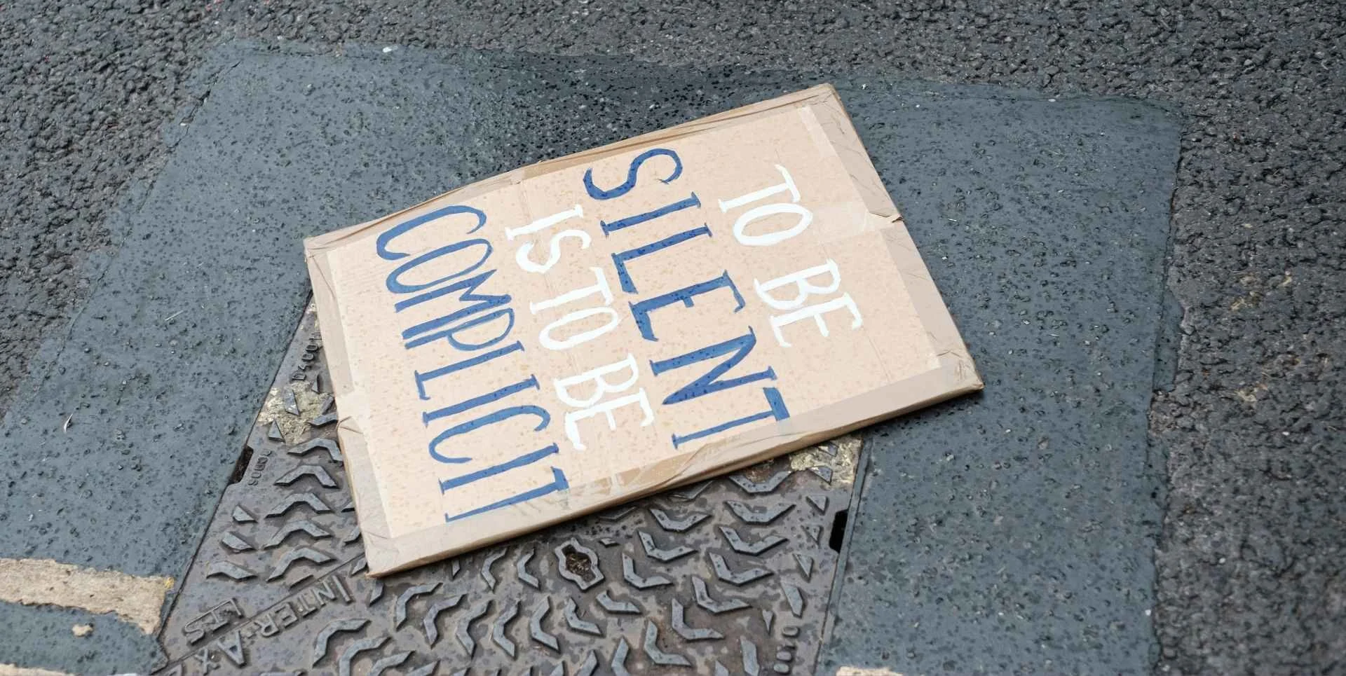 a protest sign that states to be silent is to be complicit written on cardboard and lying on the asphalt