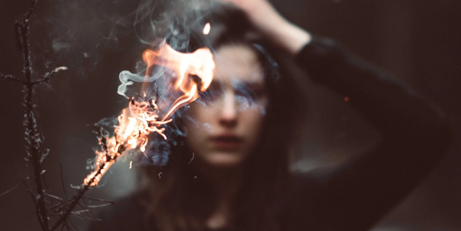 In the foreground, two bare branches, the right one is on fire. A white woman with dark hair and clothing is blurred in the background.
