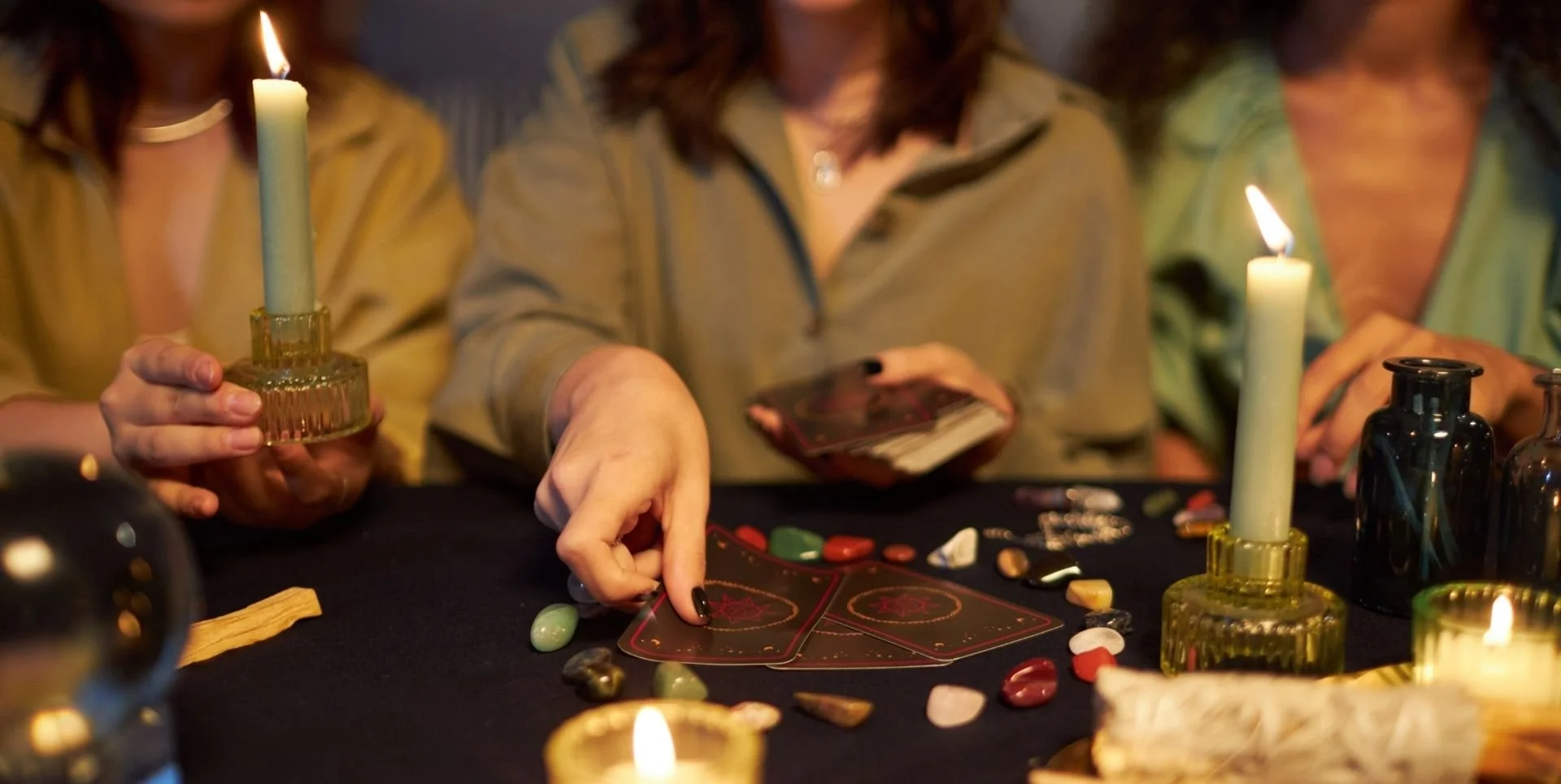 three women at a table filled with crystals and candles. The center woman is laying out tarot cards.