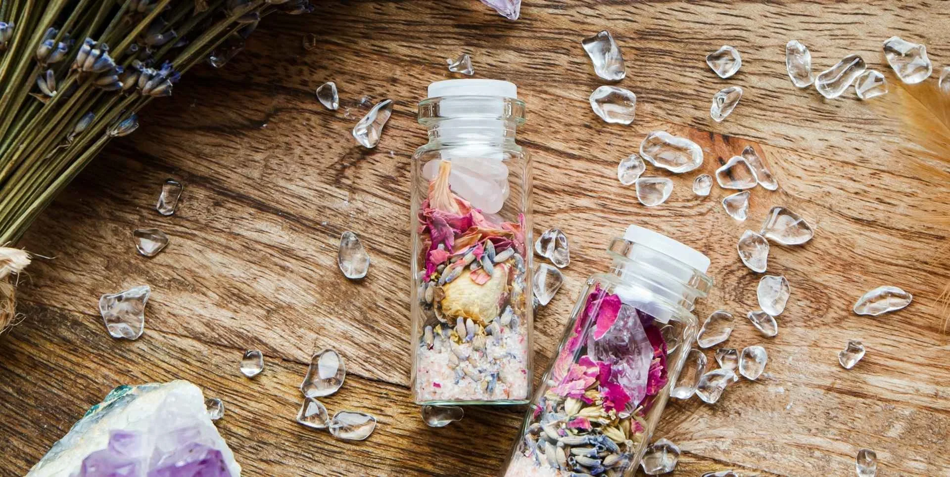 two small glass jars on a wooden table surrounded by pieces of amethyst and small clear quartz chips