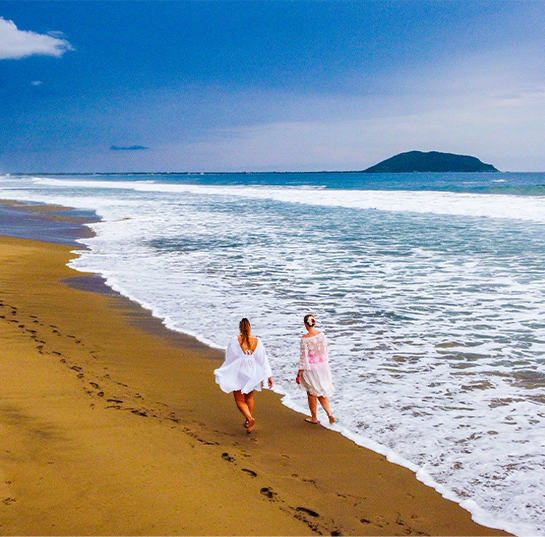 Two women walking along the sandy beach near the ocean, with footprints in the sand and an island in the distance under a partly cloudy sky.