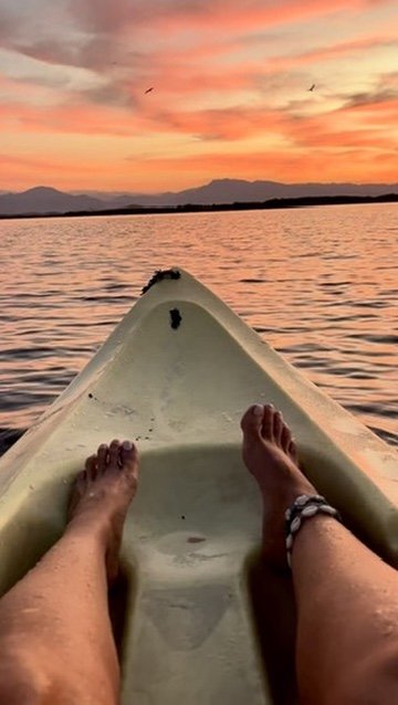 Woman kayaking on a calm river during sunset with green hills and mountains in the background.