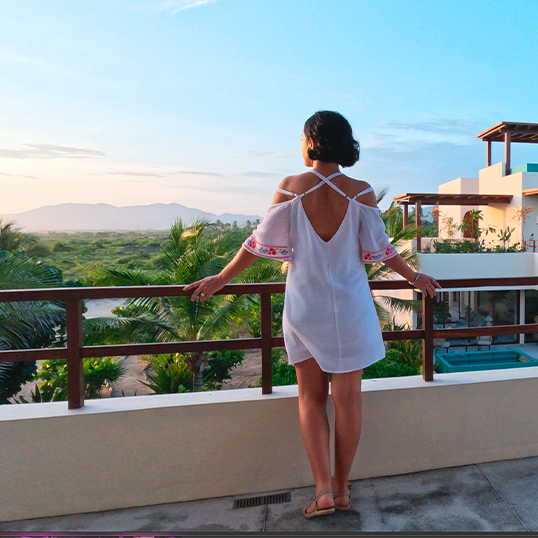 Woman standing on a balcony overlooking lush greenery and mountains at sunset.