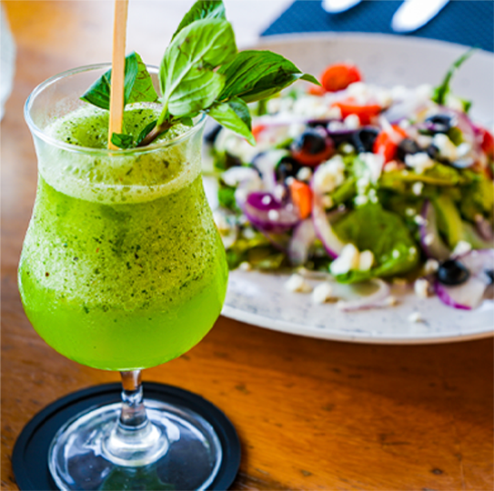 Green smoothie with basil leaves and a straw, with a colorful salad in the background on a wooden table.