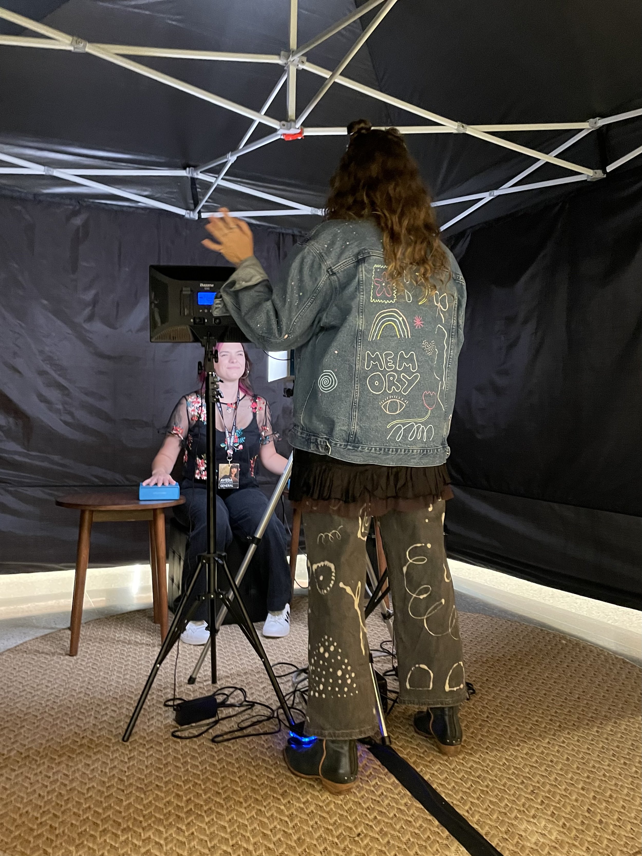 A woman sitting on a chair with a microphone and a headset, being recorded or interviewed by another woman standing with her back to the camera, inside a black soundproof booth with a fabric ceiling.